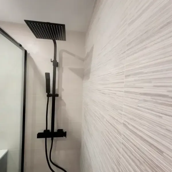 Cabezal de ducha y grifo de color negro montados sobre una pared de azulejos texturizados de color claro.