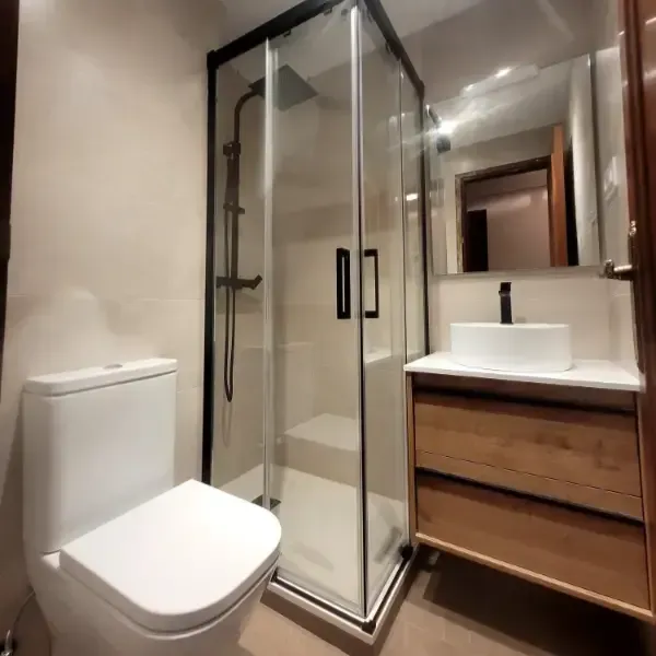 Baño moderno con inodoro blanco, ducha de cristal, tocador de madera y lavabo.