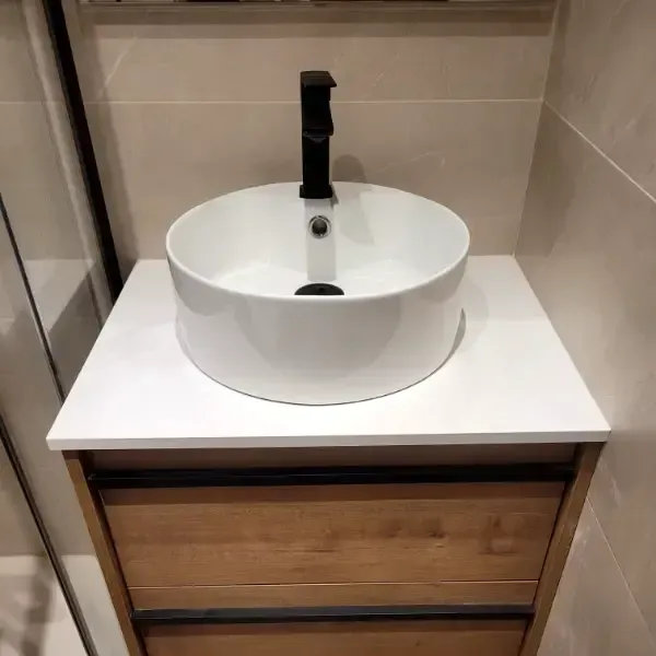 Lavabo redondo blanco sobre encimera blanca con grifo negro. Mueble de madera debajo.