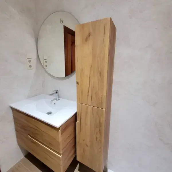 Baño con tocador de madera, mueble alto, espejo redondo y lavabo blanco.