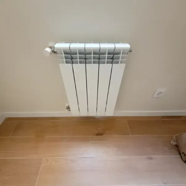 Radiador blanco montado en una pared de color beige claro sobre un piso de madera de color marrón claro.