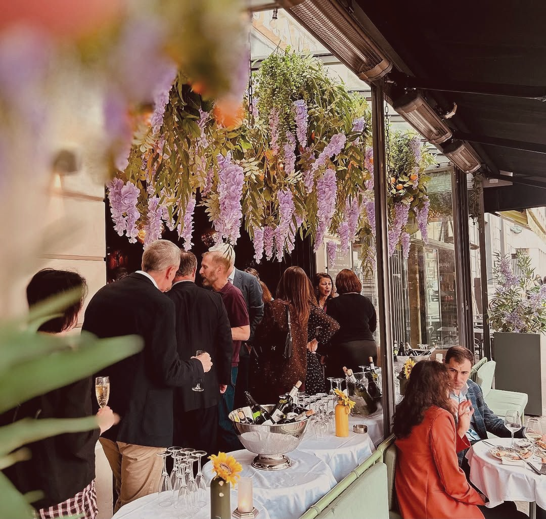 Des personnes discutent dans un restaurant en plein air décoré de glycines violettes suspendues, dans un patio couvert.