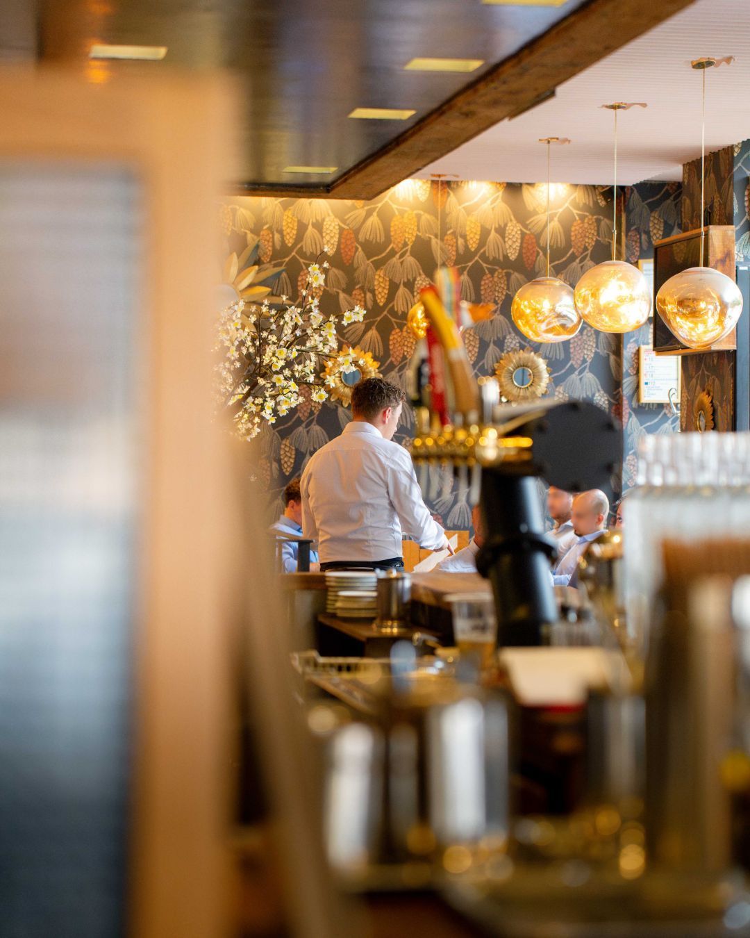 Une personne en chemise blanche se tient derrière le comptoir d'un bar dans un restaurant décoré de papier peint à motifs floraux et de suspensions en verre.
