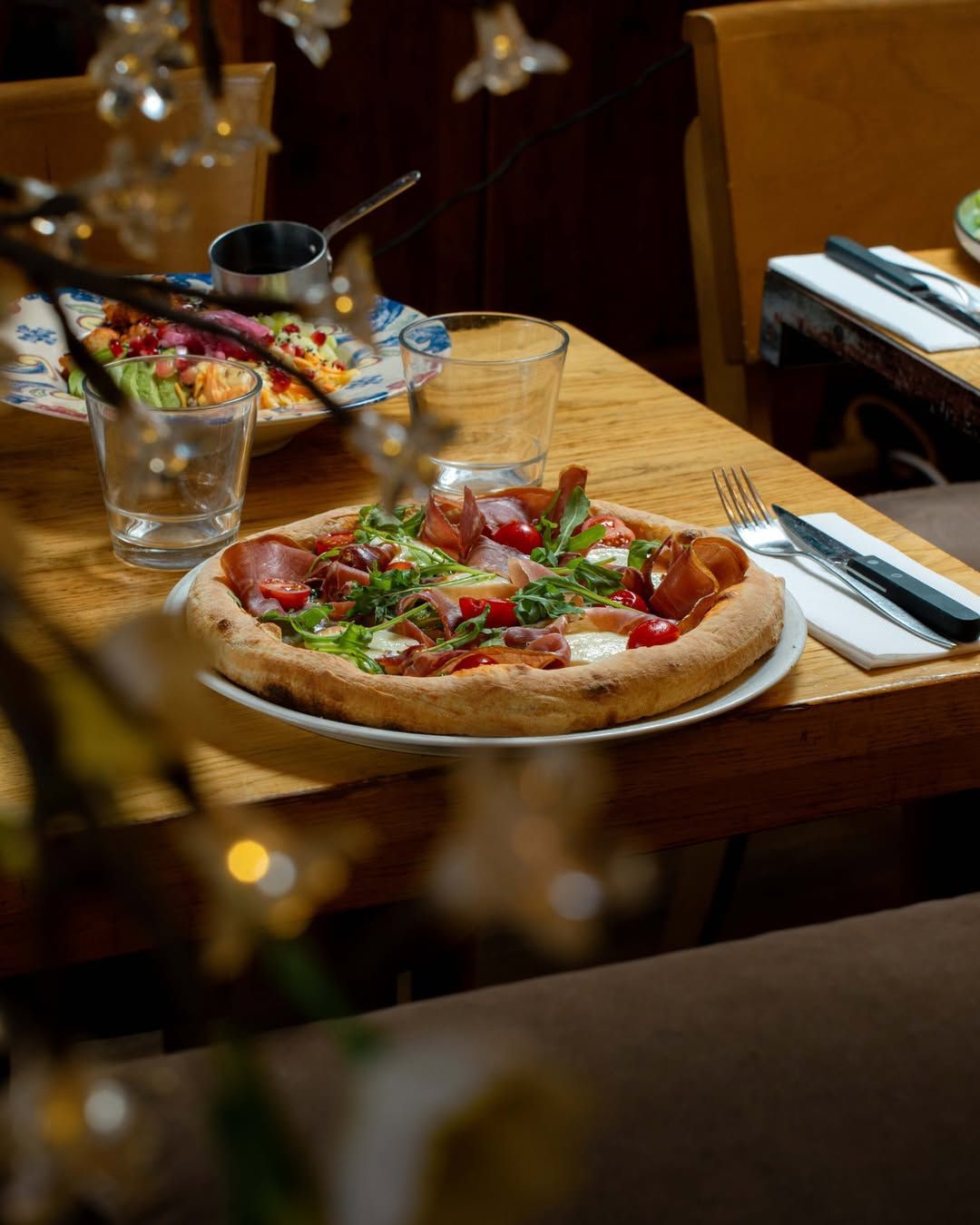 Sur une table de restaurant en bois, une pizza garnie de roquette et de prosciutto est posée à côté de couverts et d'un verre.