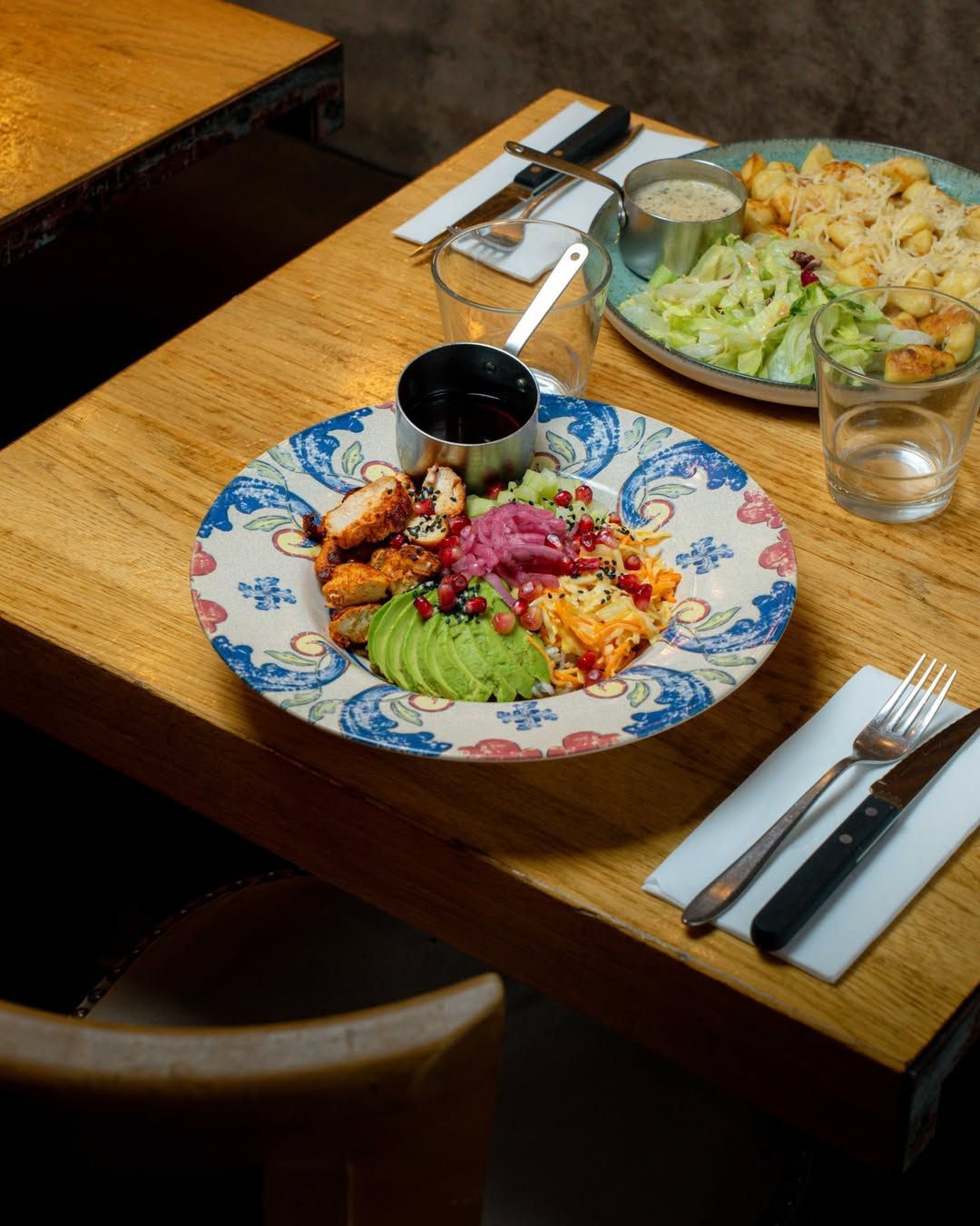 Deux bols de nourriture composés de salade, d'avocat et de viande, servis sur une table en bois clair avec des couverts et des verres.