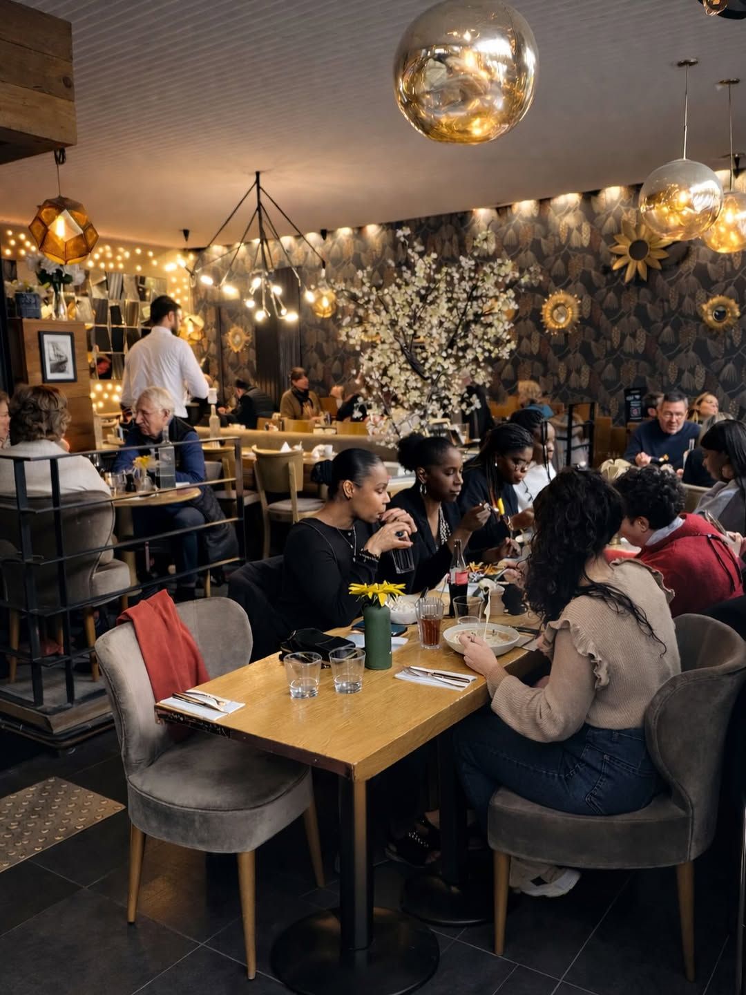 Les clients sont assis à des tables dans un restaurant moderne à l'éclairage chaleureux, décoré de luminaires sphériques dorés et de touches florales.