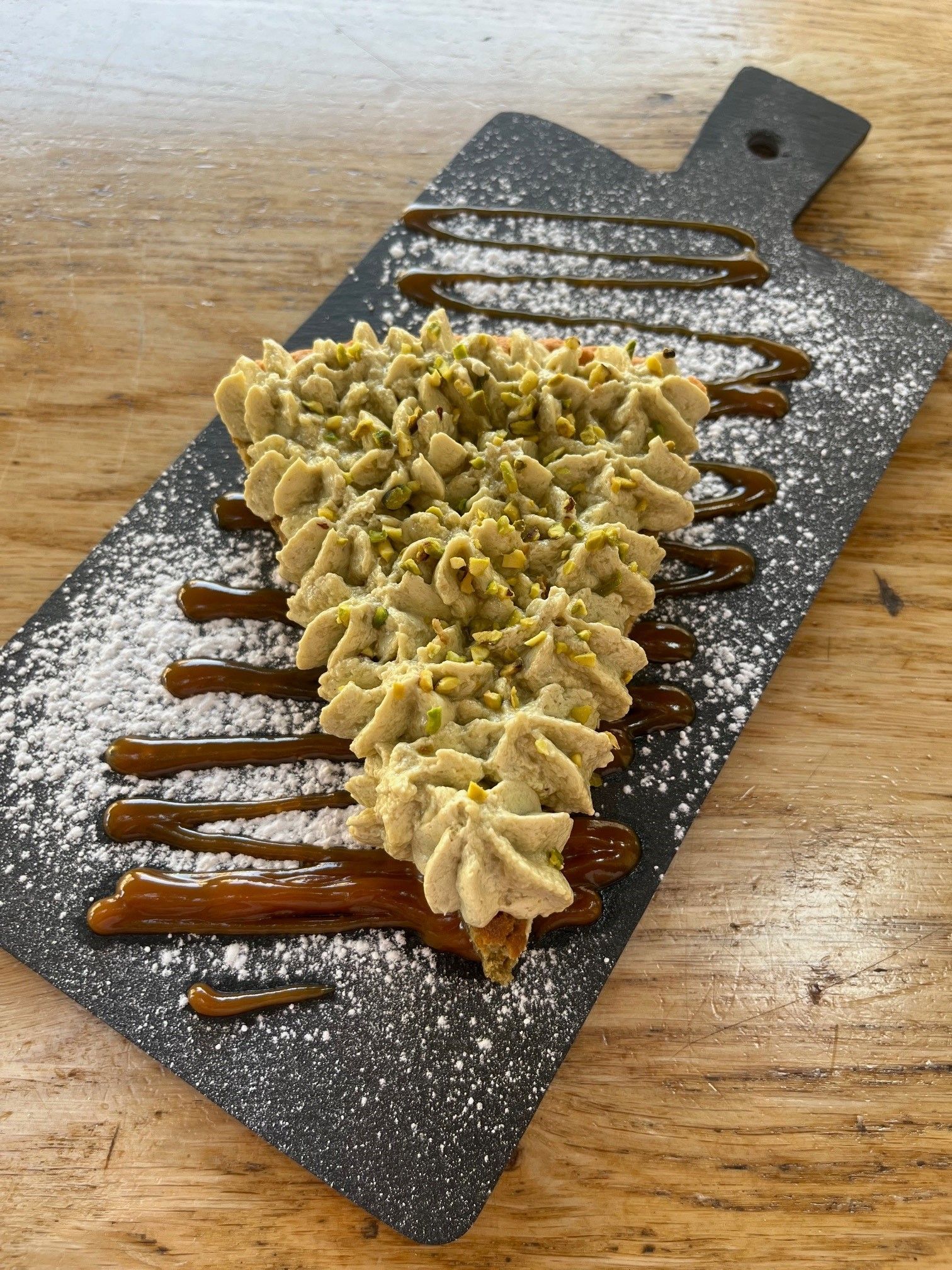 Un morceau de gâteau triangulaire garni de glaçage à la poche à douille et de pistaches concassées, arrosé de caramel et présenté sur une ardoise.