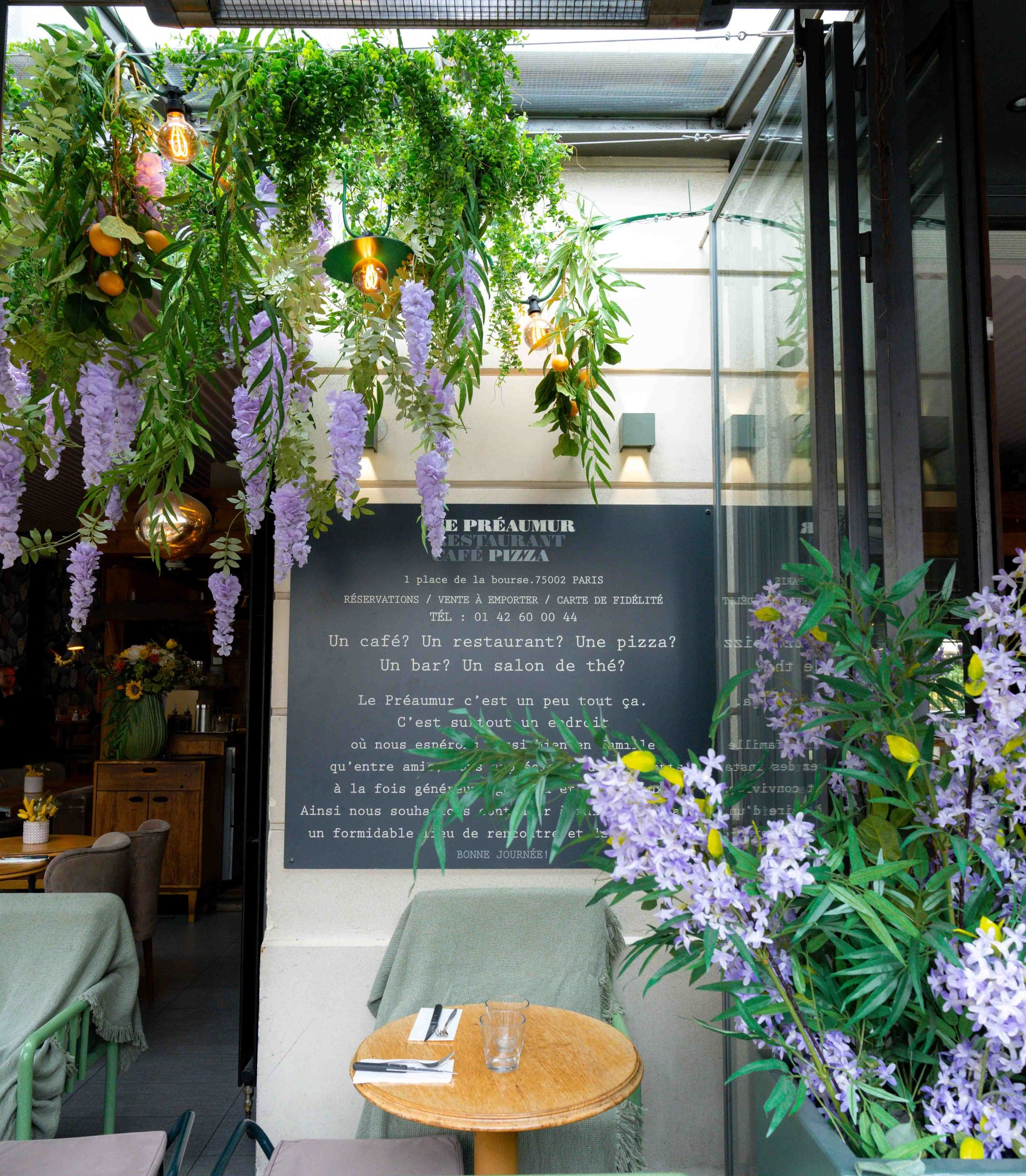 Une terrasse de café ornée de glycines violettes suspendues, un menu écrit à la craie sur un tableau noir et une petite table ronde avec des chaises.