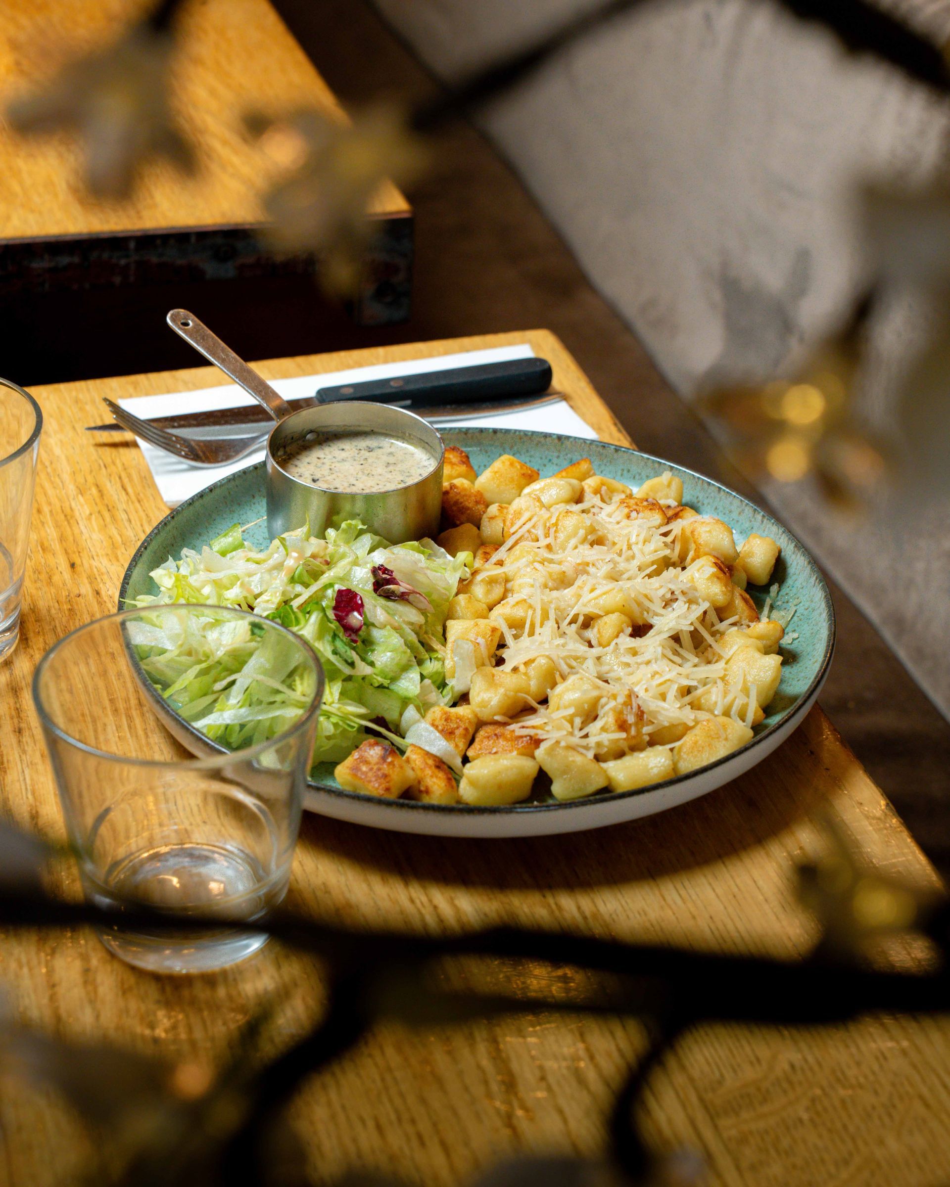 Une assiette de gnocchis gratinés au fromage, accompagnée d'une salade et servie avec une petite coupelle de sauce sur une table en bois.