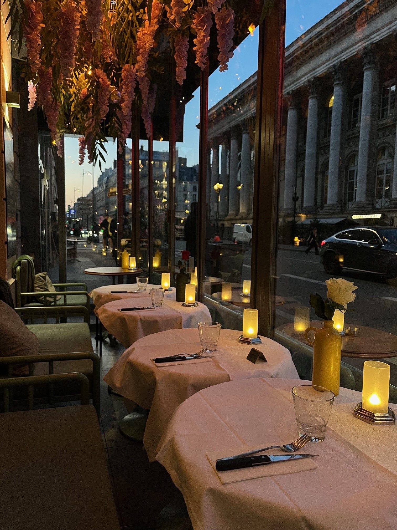 Une terrasse de restaurant avec des tables recouvertes de nappes blanches et des bougies scintillantes, donnant sur une rue au crépuscule.