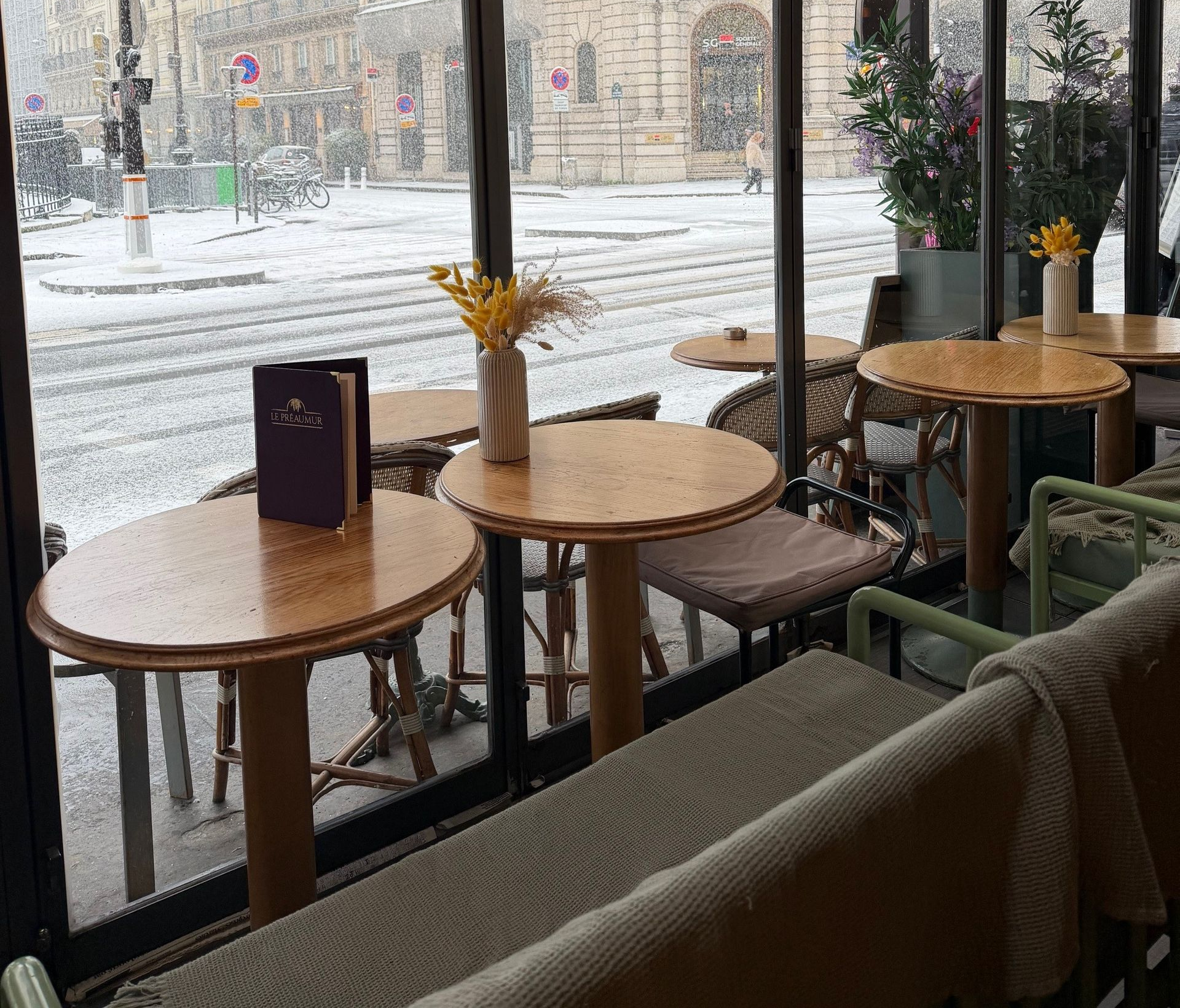 Vue depuis la fenêtre d'un café avec des tables et des chaises en bois donnant sur une rue enneigée.