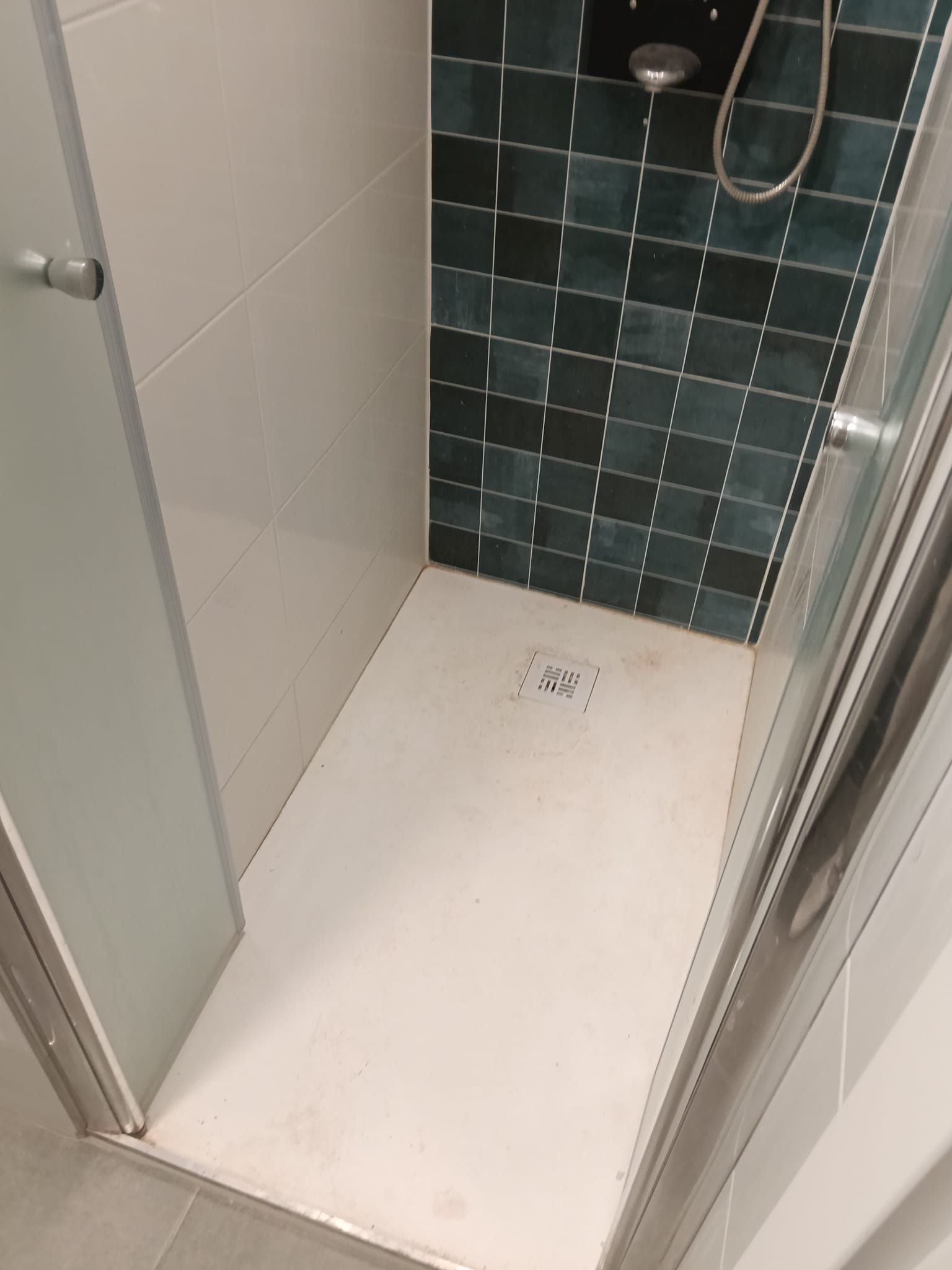 Pequeña ducha alicatada con suelo blanco, azulejos verde oscuro en la pared del fondo y una alcachofa de ducha de mano.