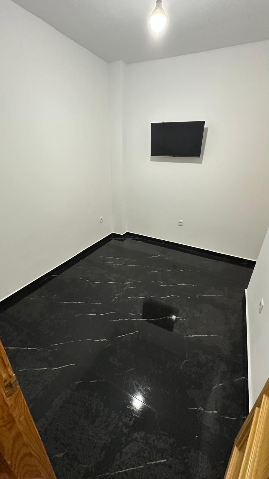 Habitación blanca vacía con suelo de baldosas negras brillantes y un televisor montado en la pared.