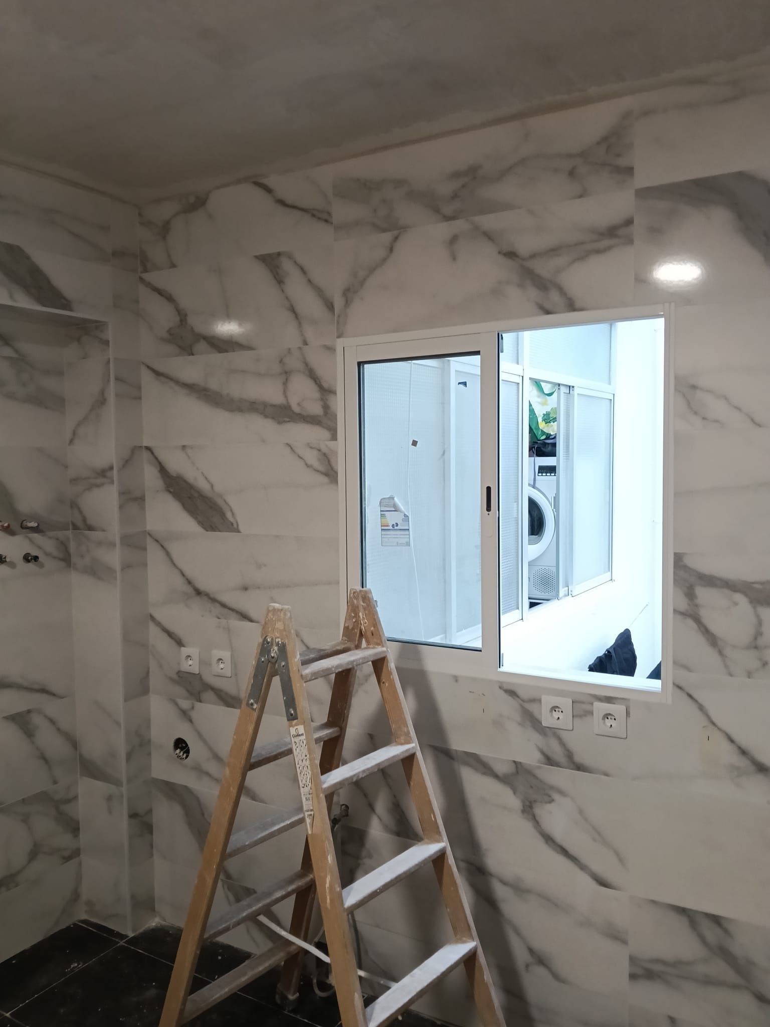 Baño revestido de mármol en proceso de reforma, con una escalera y una ventana abierta.