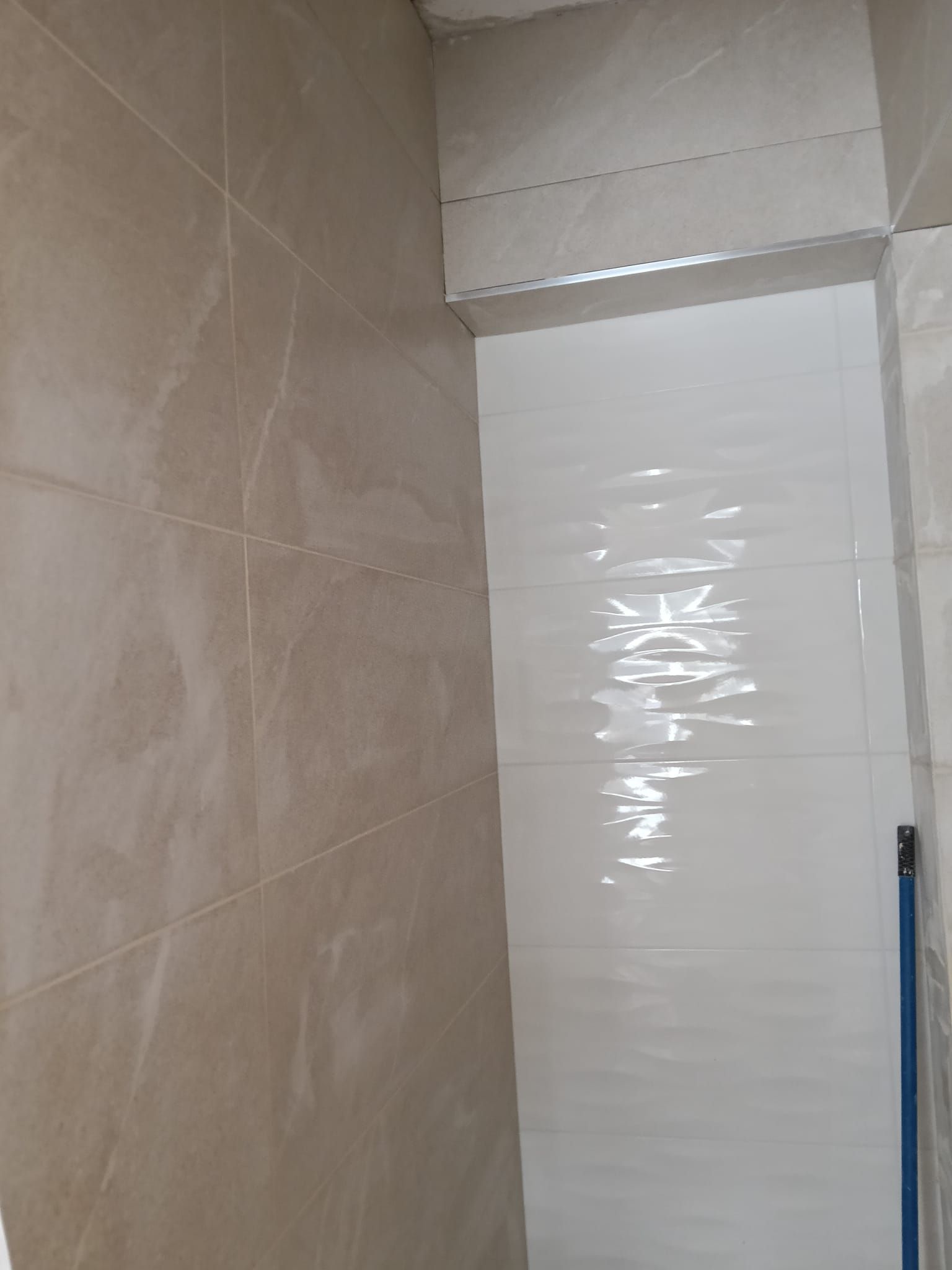 Rincón del baño con azulejos beige y un panel blanco en la pared, cerca del techo; se ve un tirador azul a la derecha.
