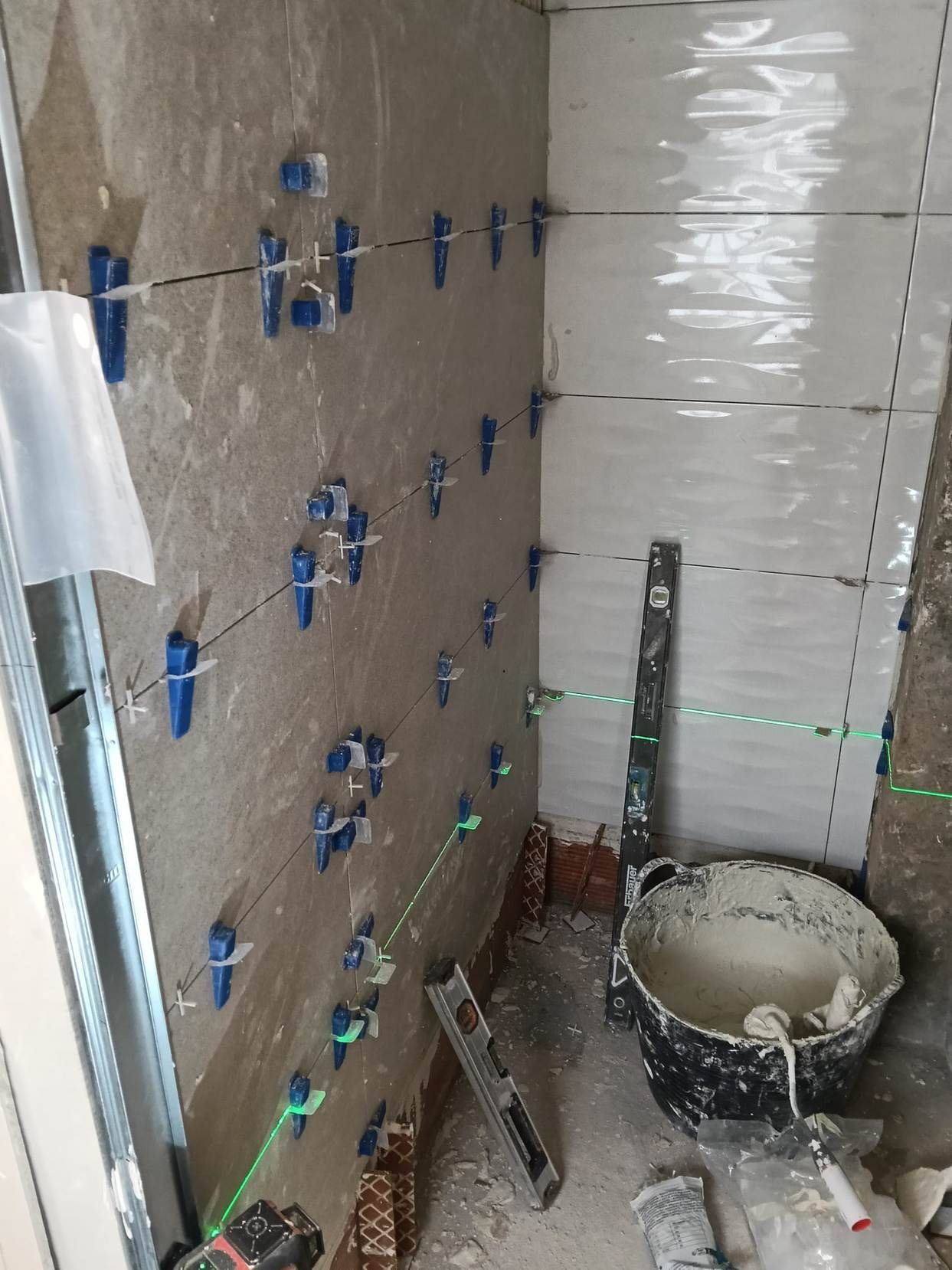 Pared parcialmente alicatada en construcción, con crucetas azules para azulejos, líneas láser verdes y un cubo en el suelo