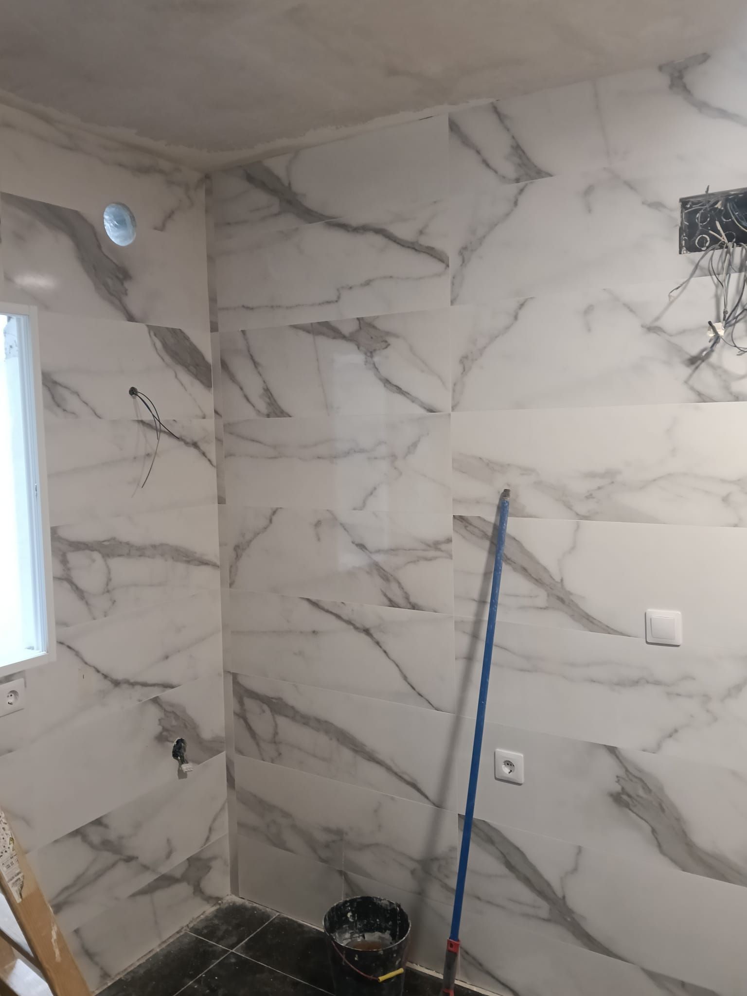 Baño pequeño con azulejos en plena renovación, con revestimiento de pared efecto mármol, ventana, enchufes, fregona y cubo.
