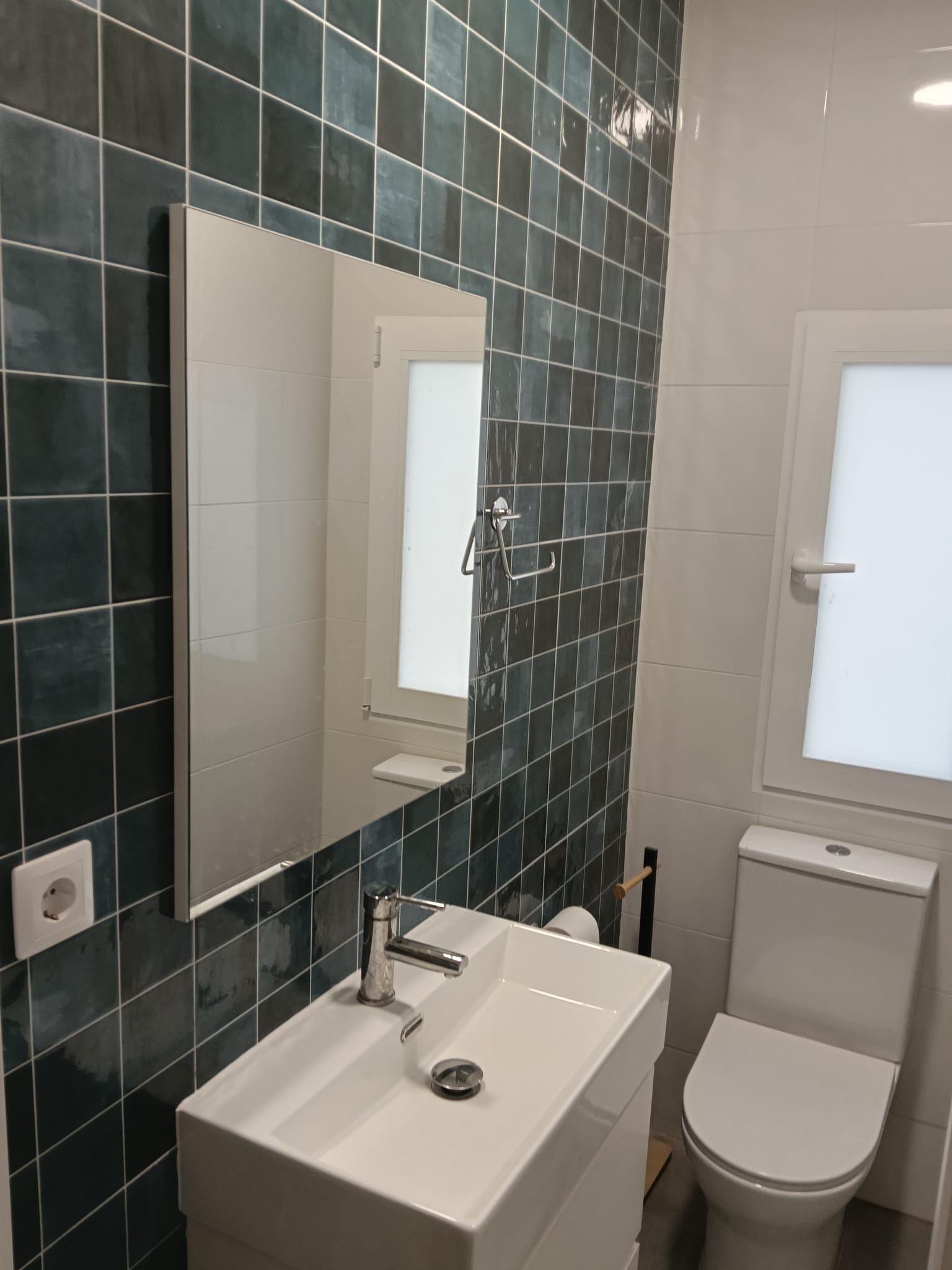Baño moderno con una pared revestida de azulejos verde oscuro, espejo, lavabo, inodoro y ventana esmerilada.