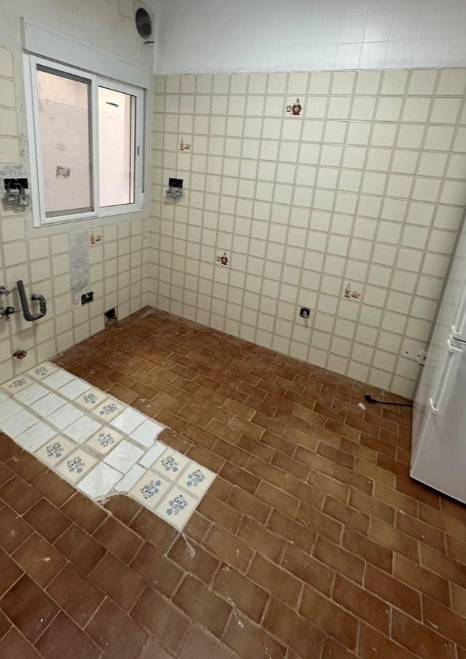 Baño vacío con suelo marrón y paredes blancas alicatadas, con una zona de ducha y una ventana.