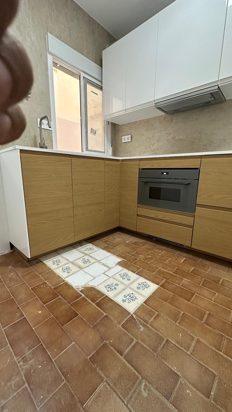 Cocina pequeña con armarios de madera, horno empotrado, suelo de terracota y una alfombrilla de azulejos blancos junto a la encimera.