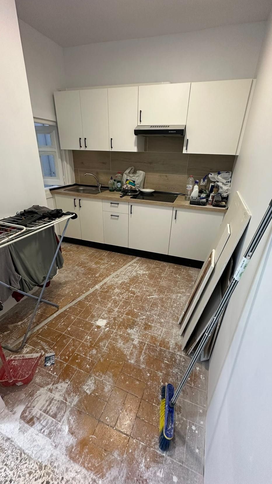 Cocina pequeña con armarios blancos, una encimera marrón y productos de limpieza en el suelo desordenado.