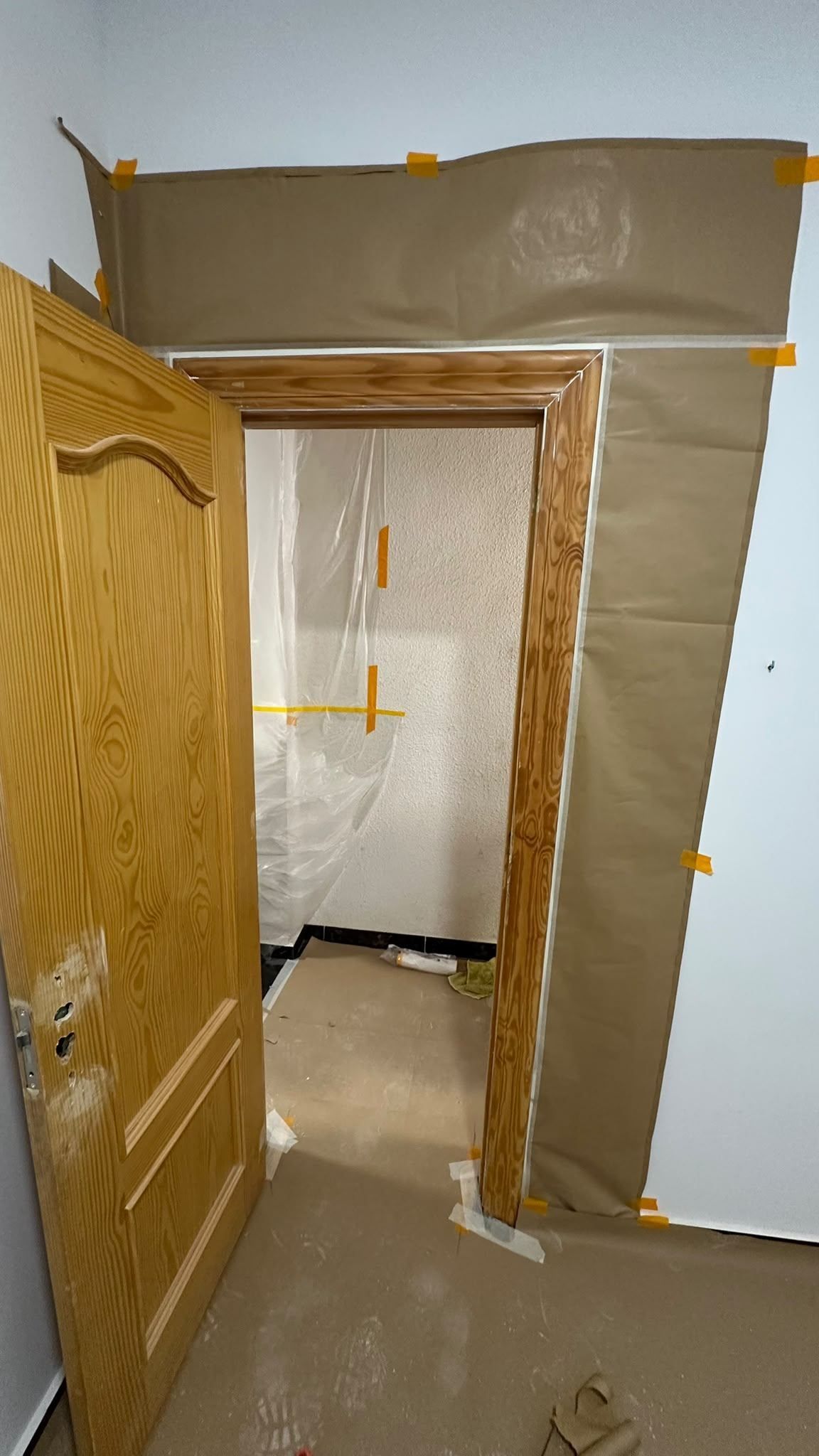 Entrada en obras, con plástico protector sujeto con cinta y placas de yeso alrededor del marco de una puerta de madera.