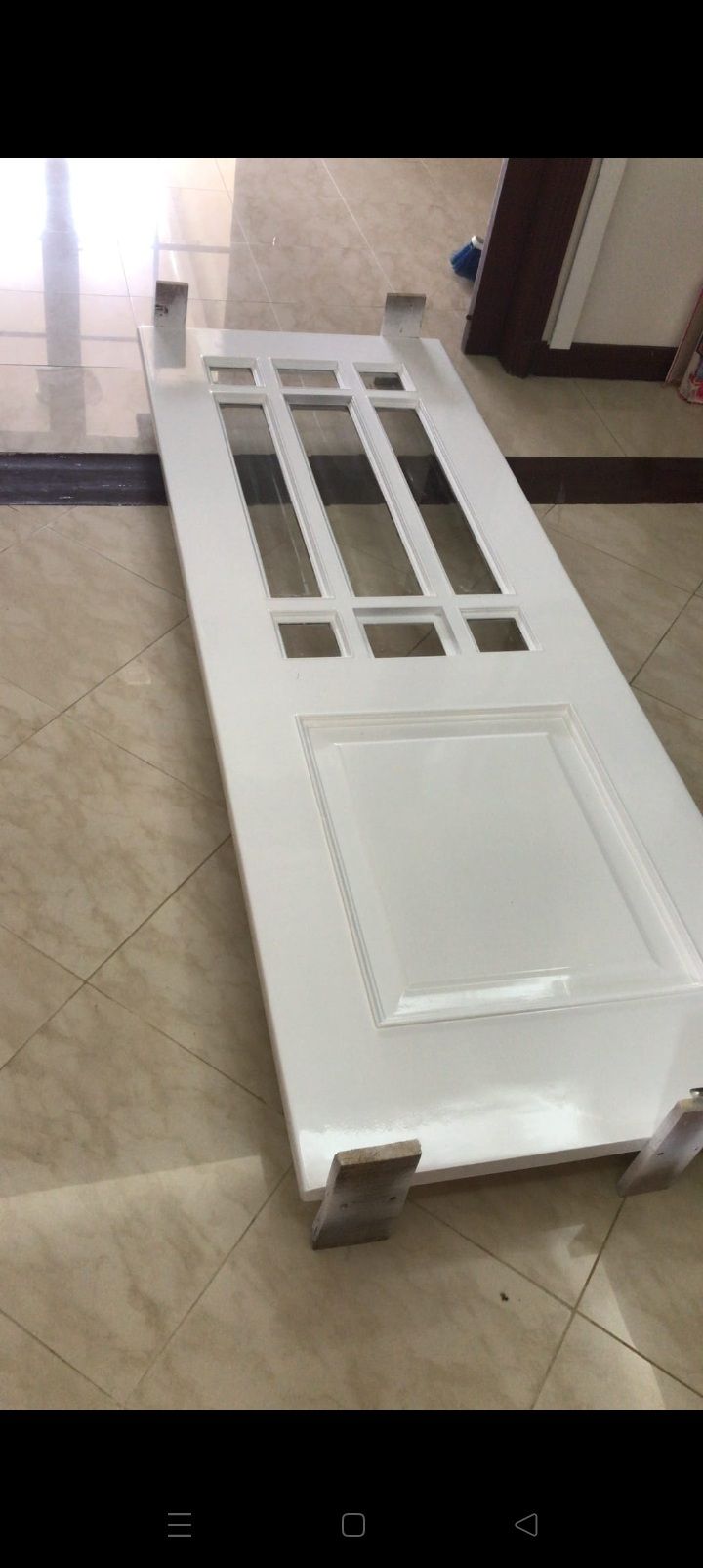 Puerta blanca de interior tumbada sobre un suelo de baldosas, vista desde arriba, con huecos para paneles de vidrio y un panel inferior encastrado
