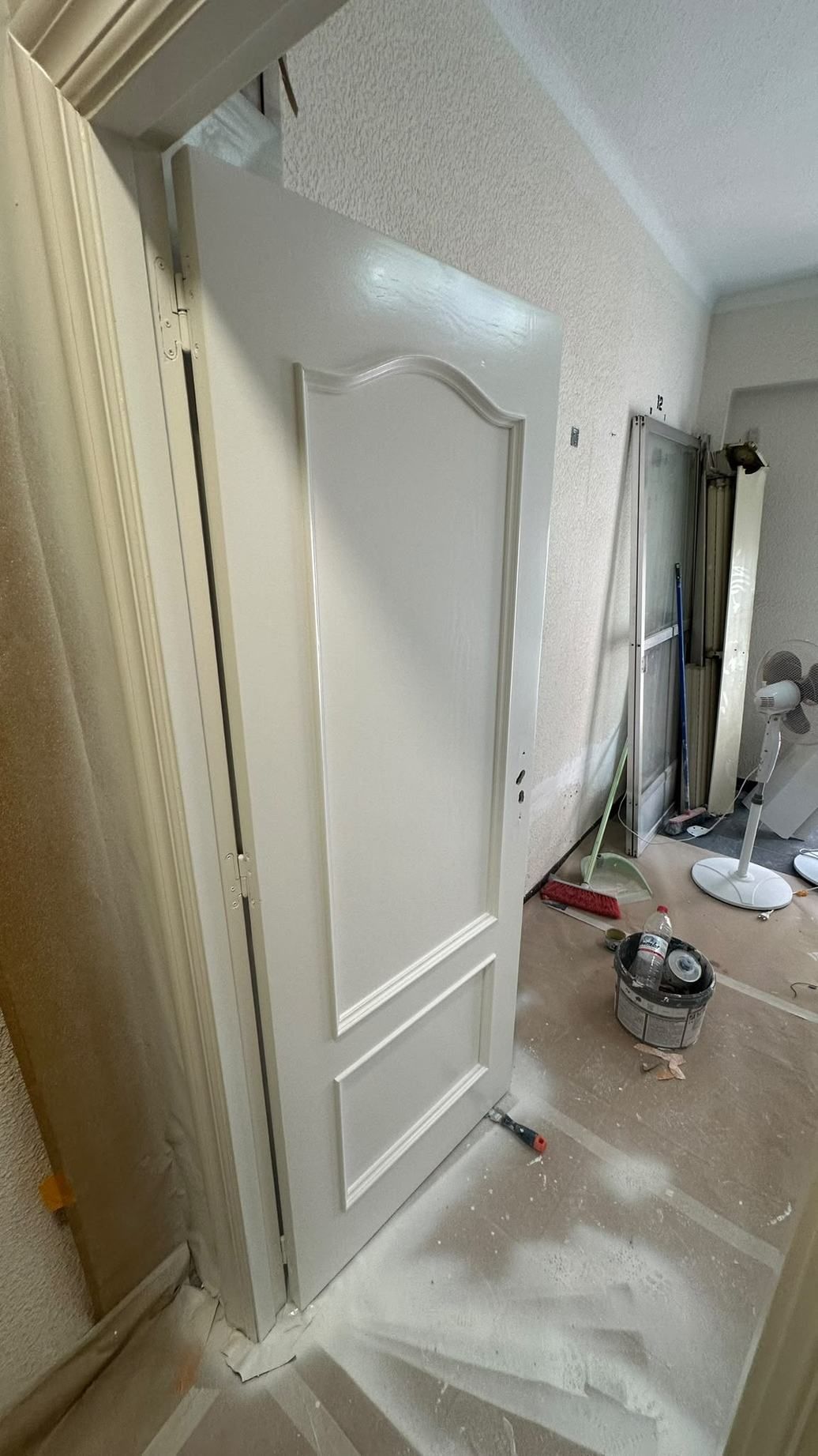 Puerta blanca con paneles apoyada en una habitación polvorienta en obras, con herramientas y materiales de pintura cerca.