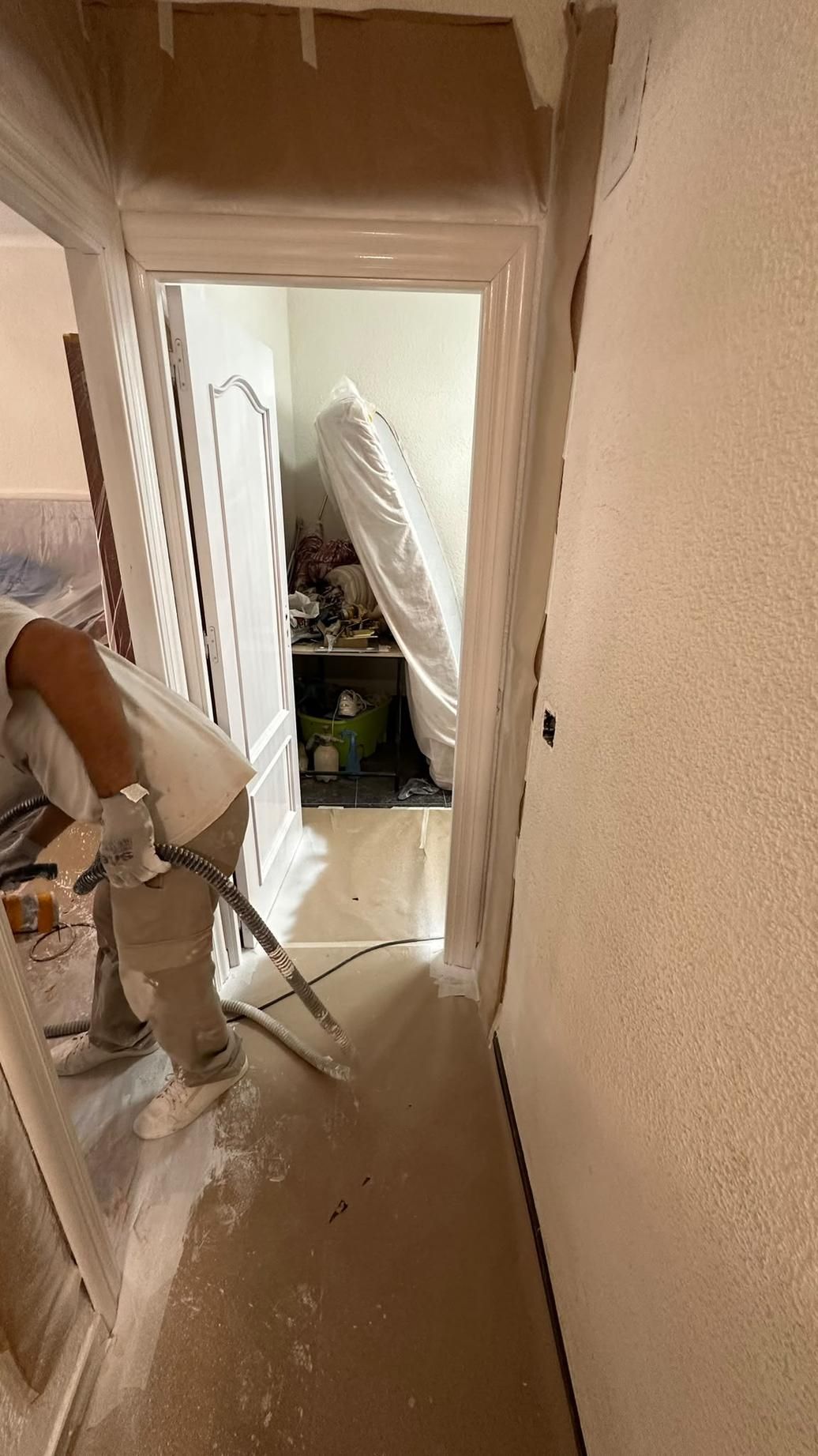 Pasillo estrecho en obras, con el suelo cubierto de polvo, herramientas de pladur y muebles cubiertos con plástico al fondo