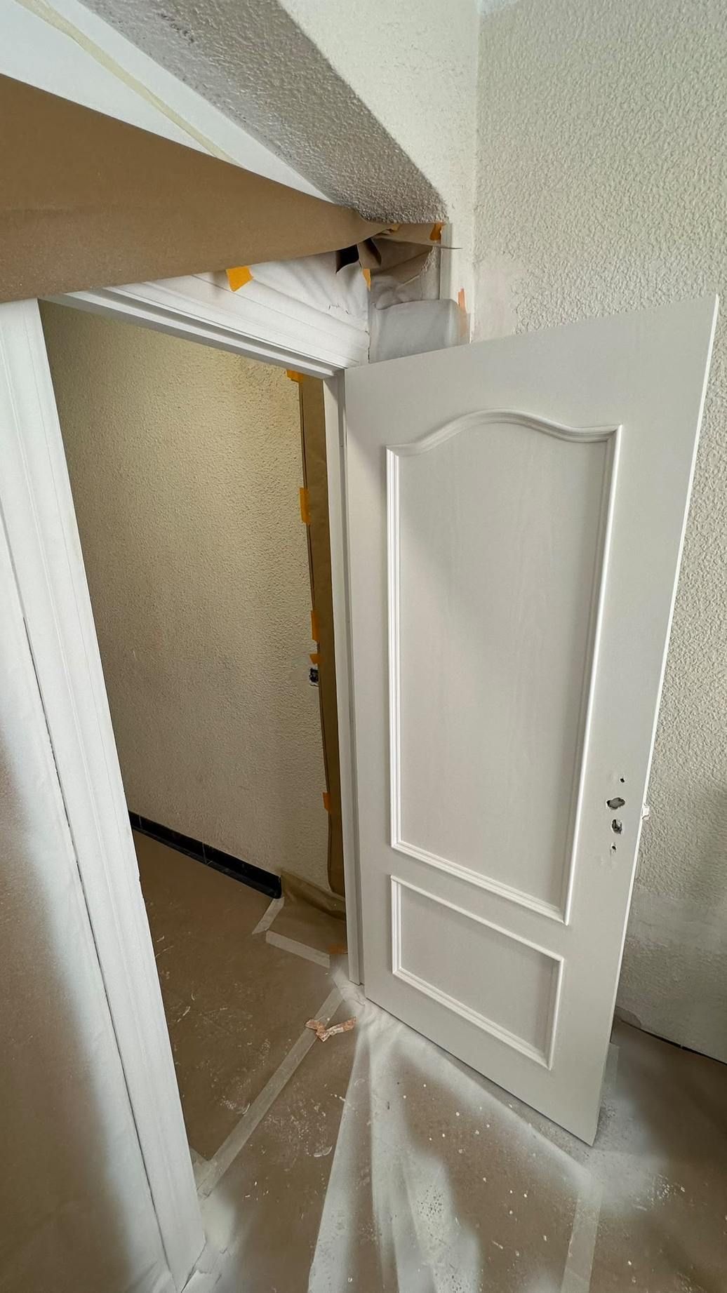 Puerta blanca entreabierta bajo una escalera en un pasillo estrecho, con escombros en el suelo.
