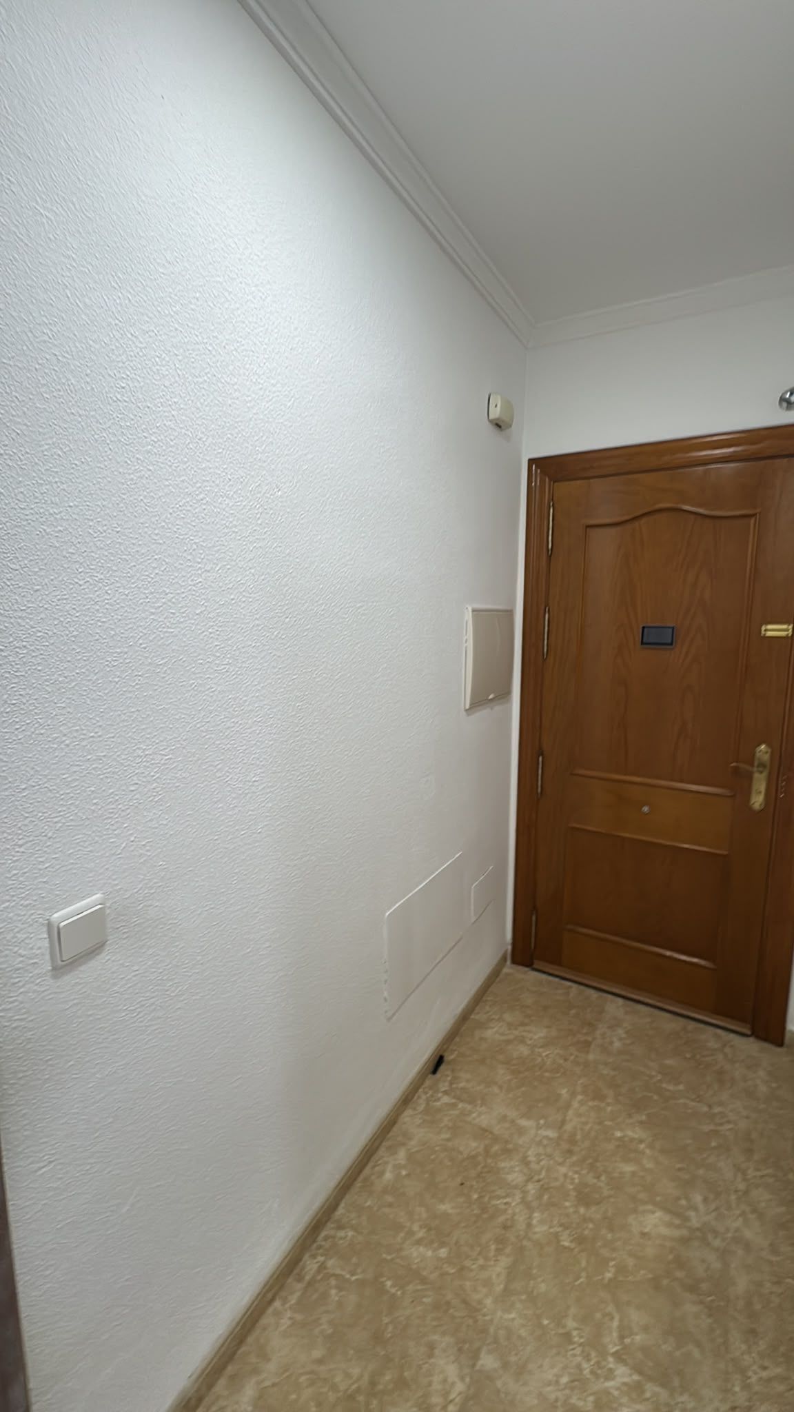 Pasillo estrecho con paredes blancas texturizadas, suelo beige y una puerta de entrada de madera