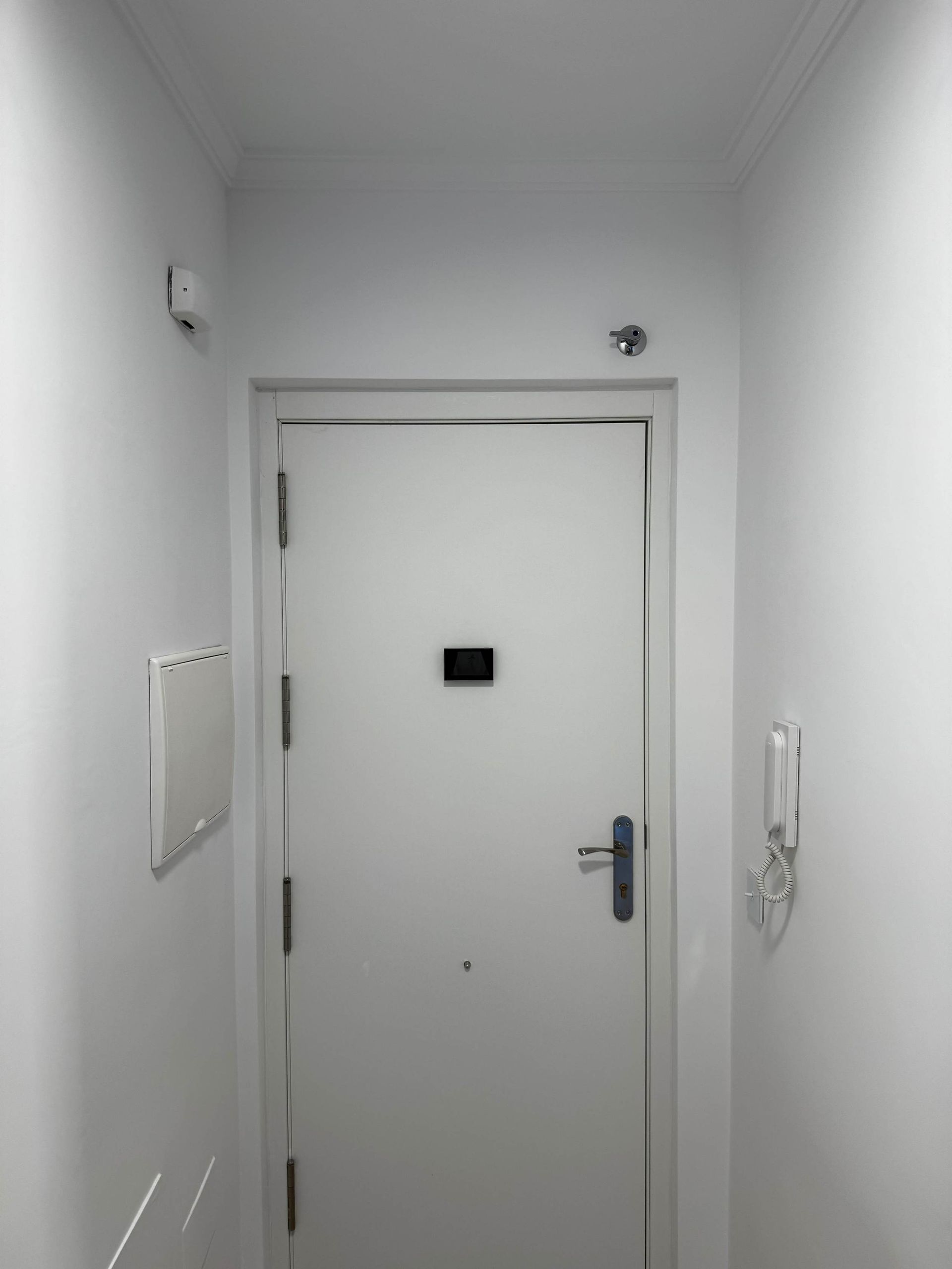 Pasillo blanco con una puerta blanca cerrada, una mirilla y un interfono montado en la pared.