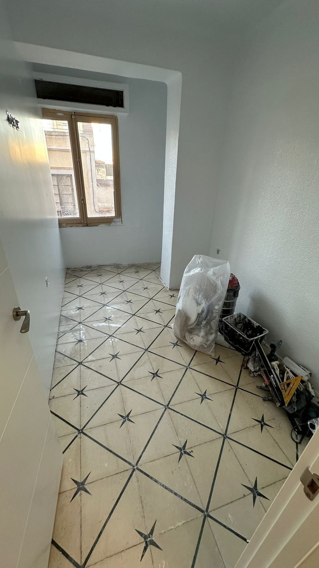Habitación vacía con suelo de baldosas, una ventana y materiales de construcción junto a la pared derecha.