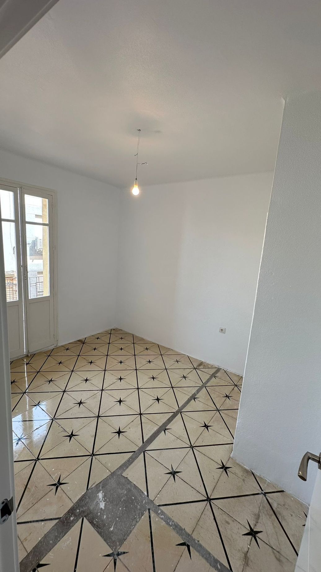 Habitación blanca vacía con suelo de baldosas estampadas, puerta con ventanas y una bombilla desnuda colgando