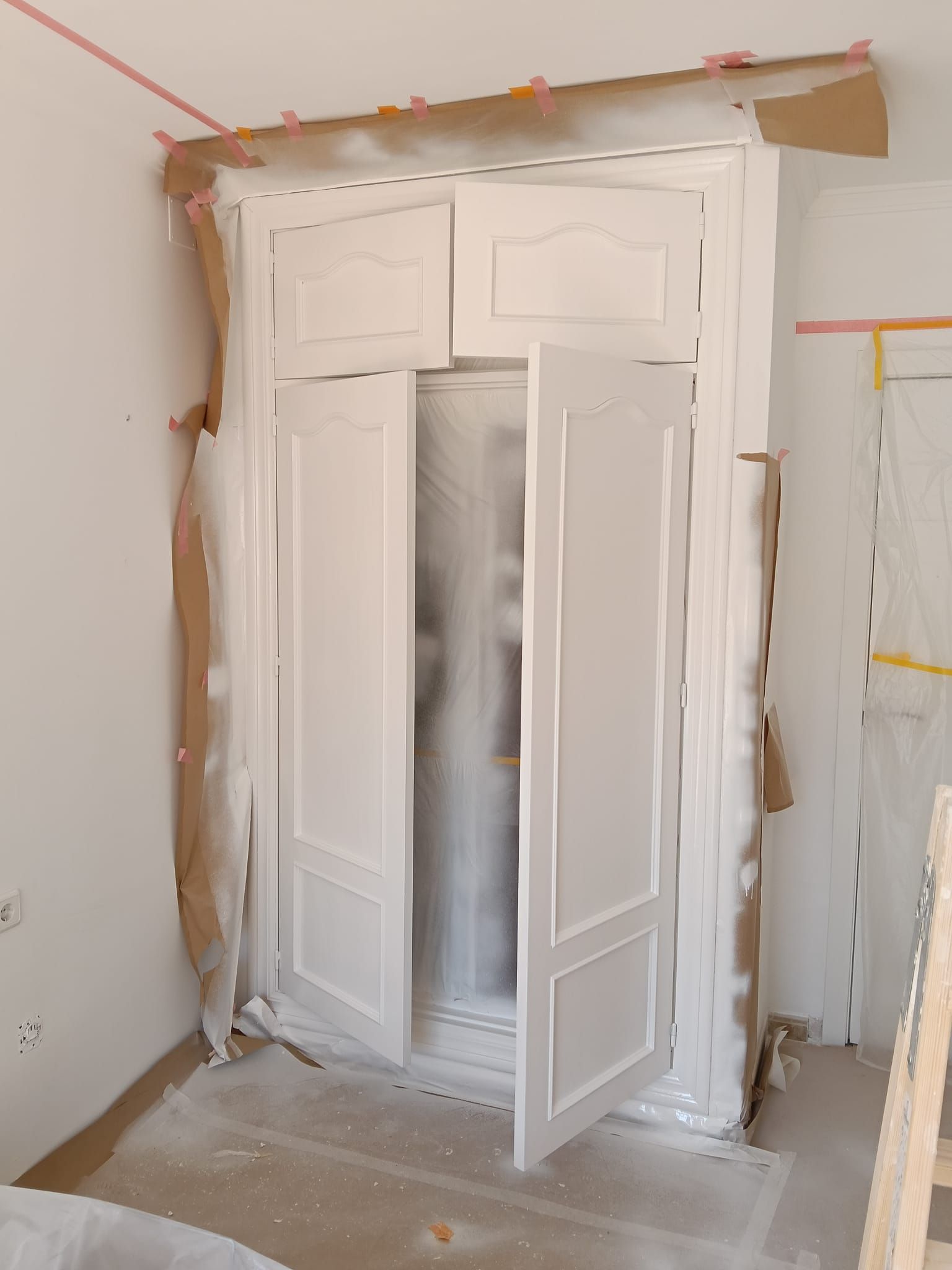 Puertas dobles blancas en una habitación en remodelación, entreabiertas entre yeso y cinta de carrocero.