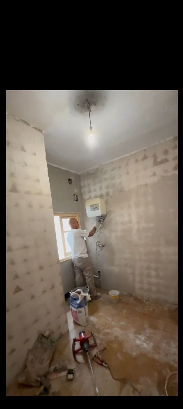 Una persona pinta una pared beige en una habitación pequeña, con herramientas y materiales en el suelo.
