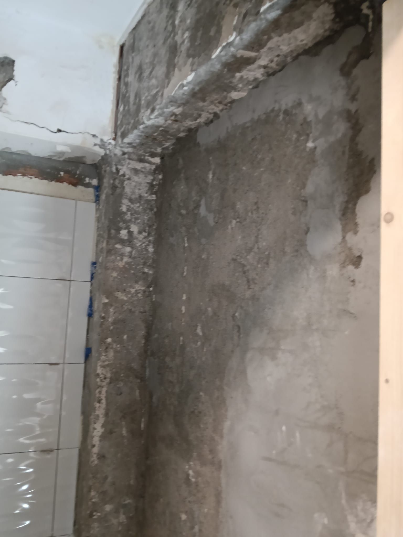 Hueco en una pared de hormigón sin terminar junto a una pared alicatada de blanco, en un baño o cuarto de servicio