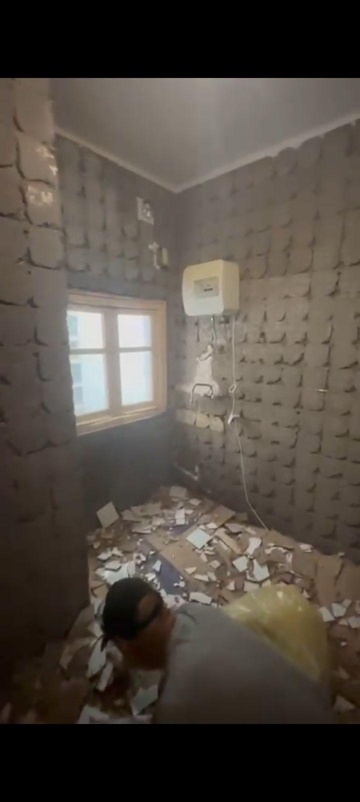 Pequeña habitación alicatada con una ventana, el suelo cubierto de escombros y una persona agachada junto a una cama.