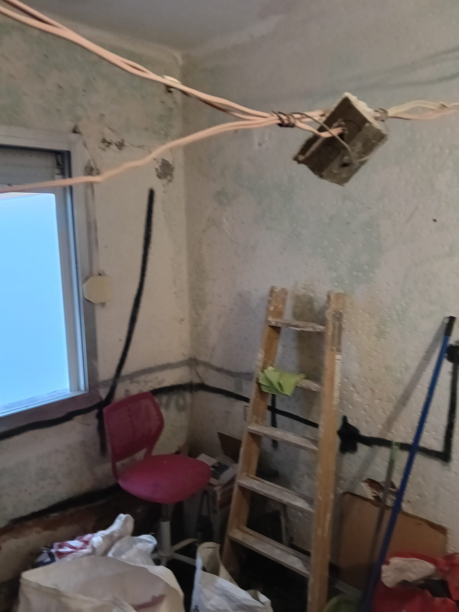 Pequeña habitación sin terminar con una escalera, una silla rosa, cables a la vista y una ventana con el marco azul.