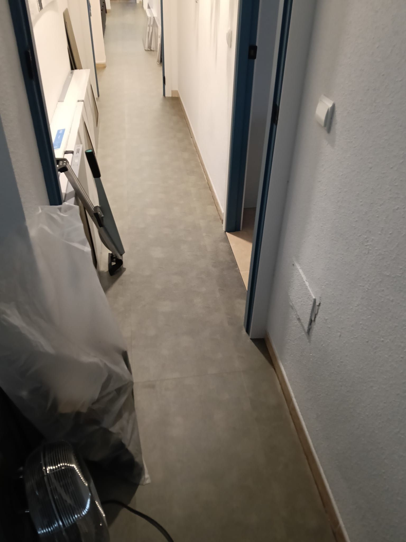Pasillo estrecho con suelo gris, paredes blancas y una puerta con molduras azules; hay objetos desordenados a lo largo del lado izquierdo.