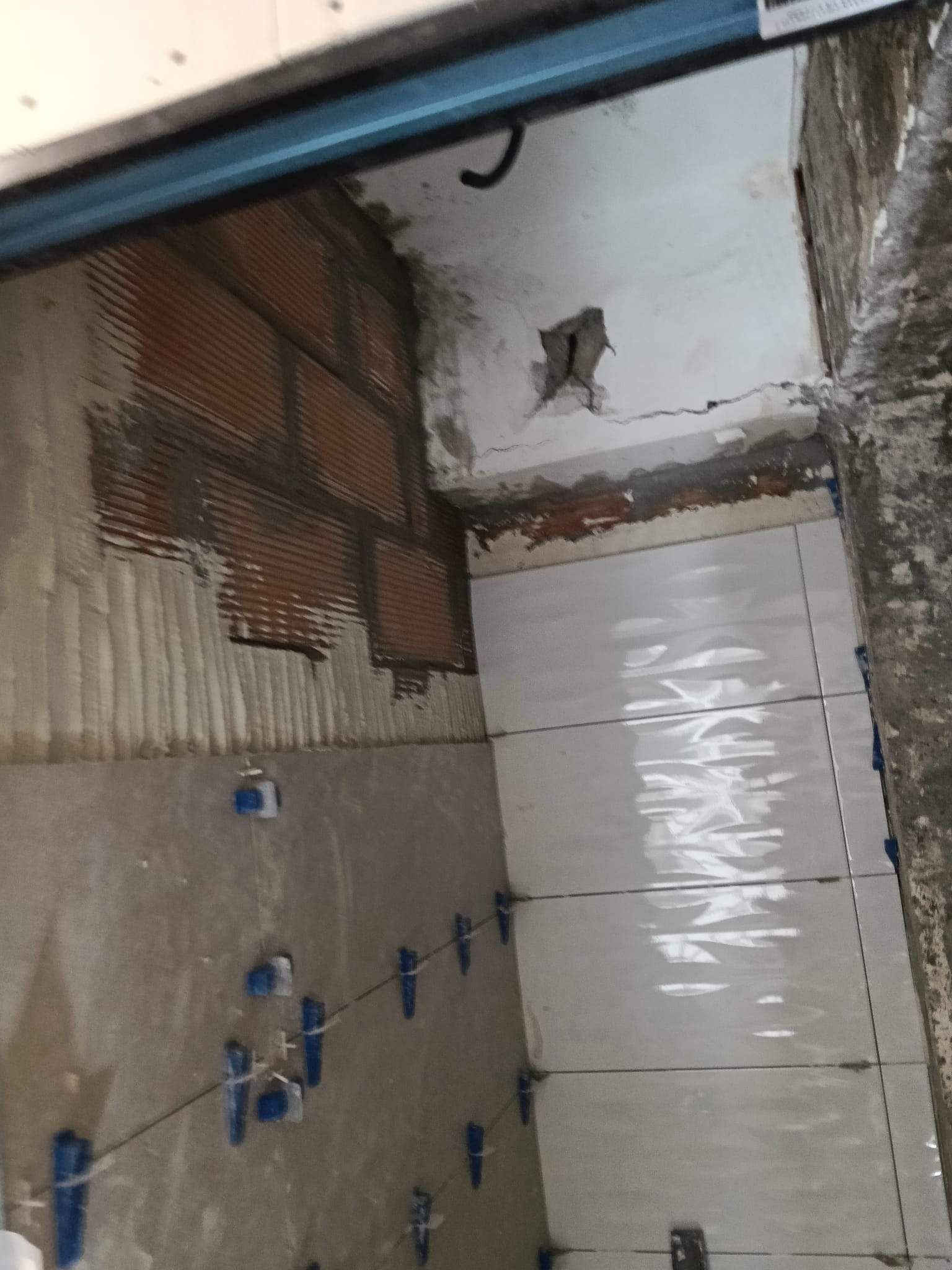 Baño pequeño sin terminar, con las paredes al descubierto, azulejos blancos, tuberías azules y una abertura en el techo