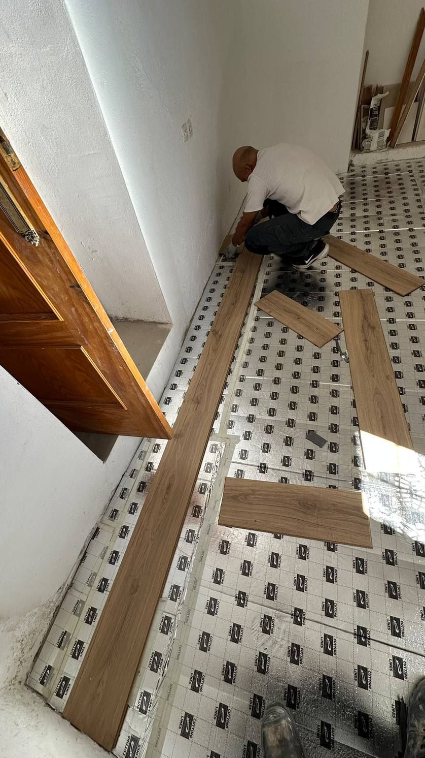 Una persona arrodillada en el suelo de un pasillo, trabajando junto a tablas de madera y herramientas esparcidas.