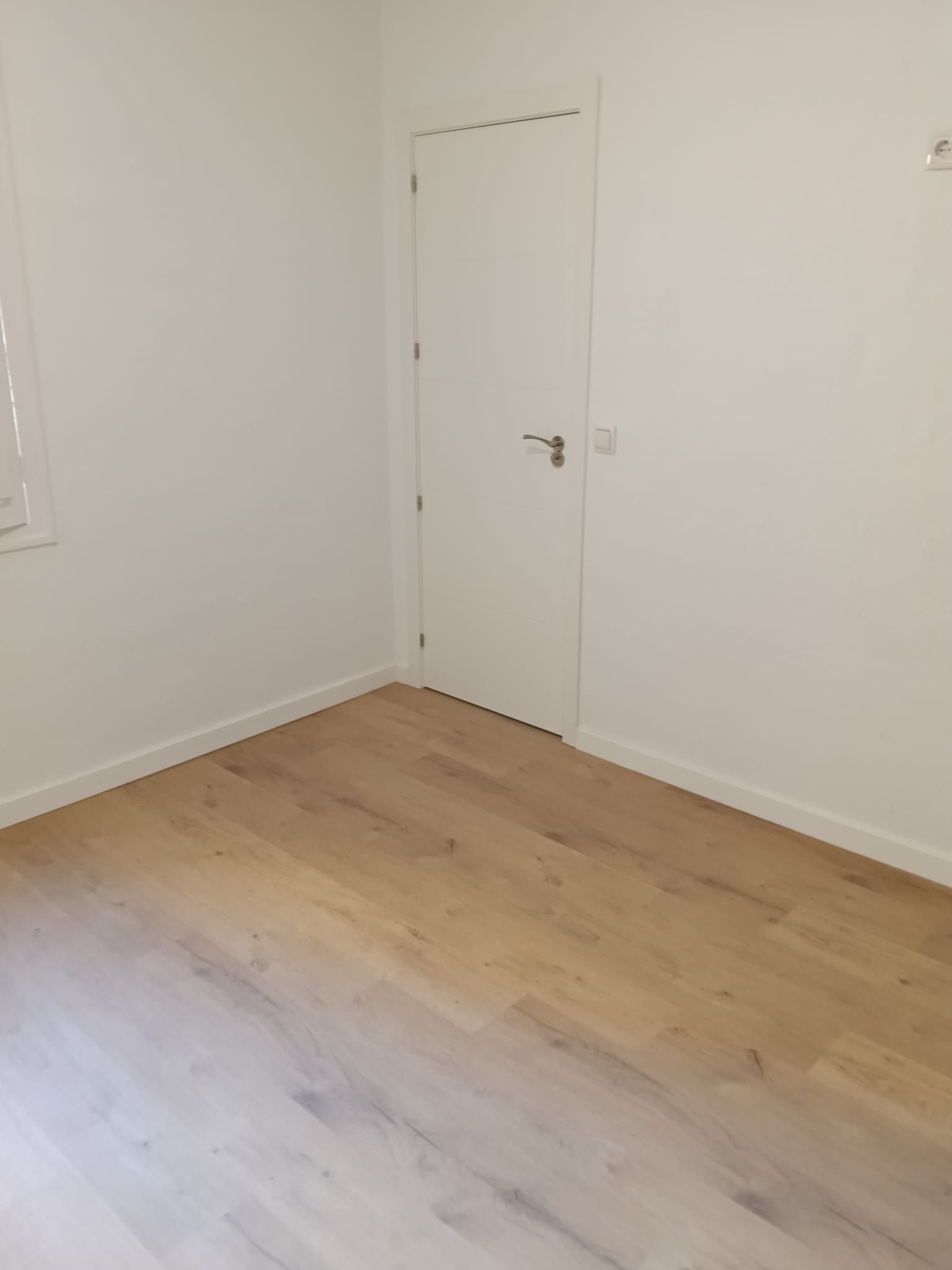 Habitación blanca y vacía, con suelo de madera clara y una puerta cerrada