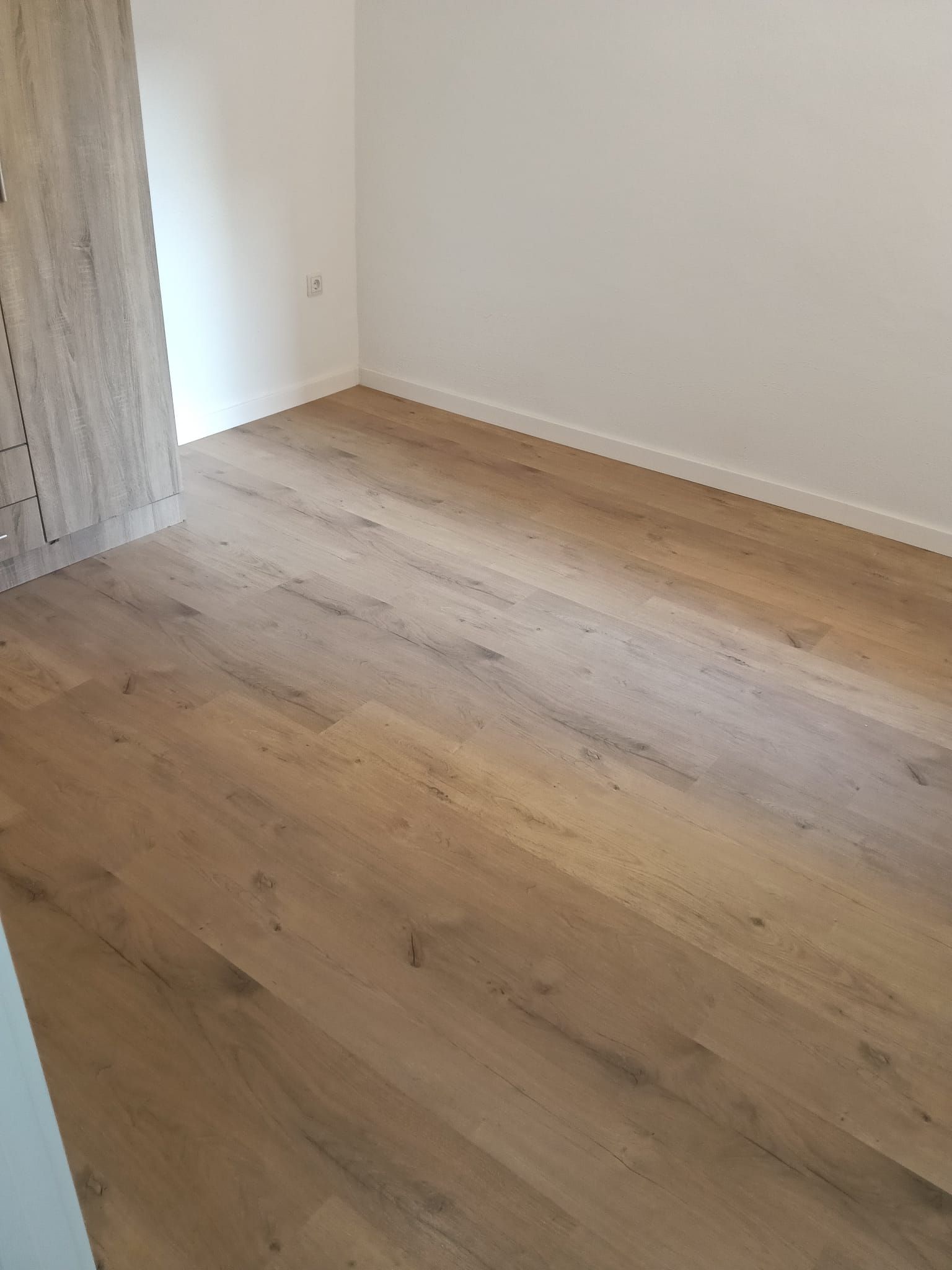 Habitación vacía con suelo de madera clara, paredes blancas y una puerta a la izquierda.