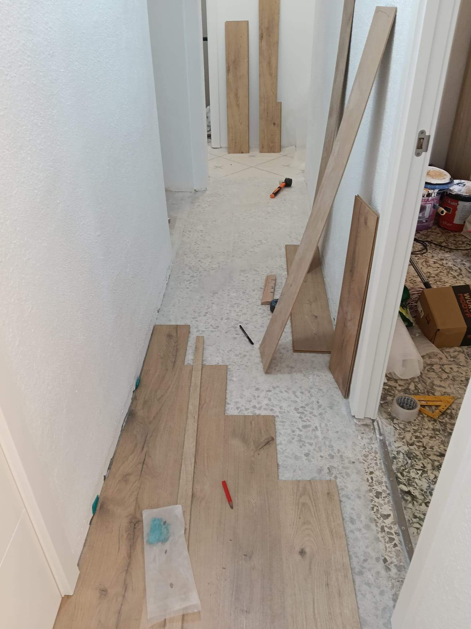 Pasillo en obras, con tablones de madera, herramientas y escombros en el suelo, además de unas tablas apoyadas junto a una puerta