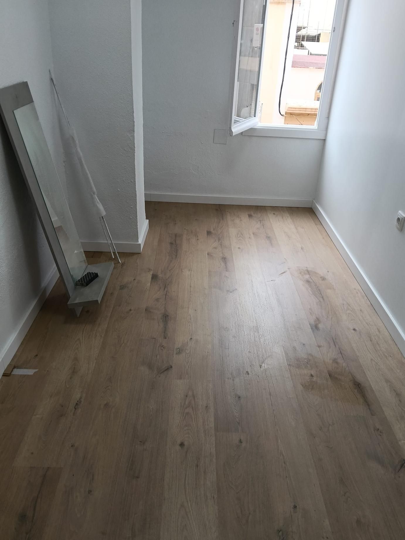 Habitación estrecha y vacía, con suelo de madera, paredes blancas y una ventana al fondo.