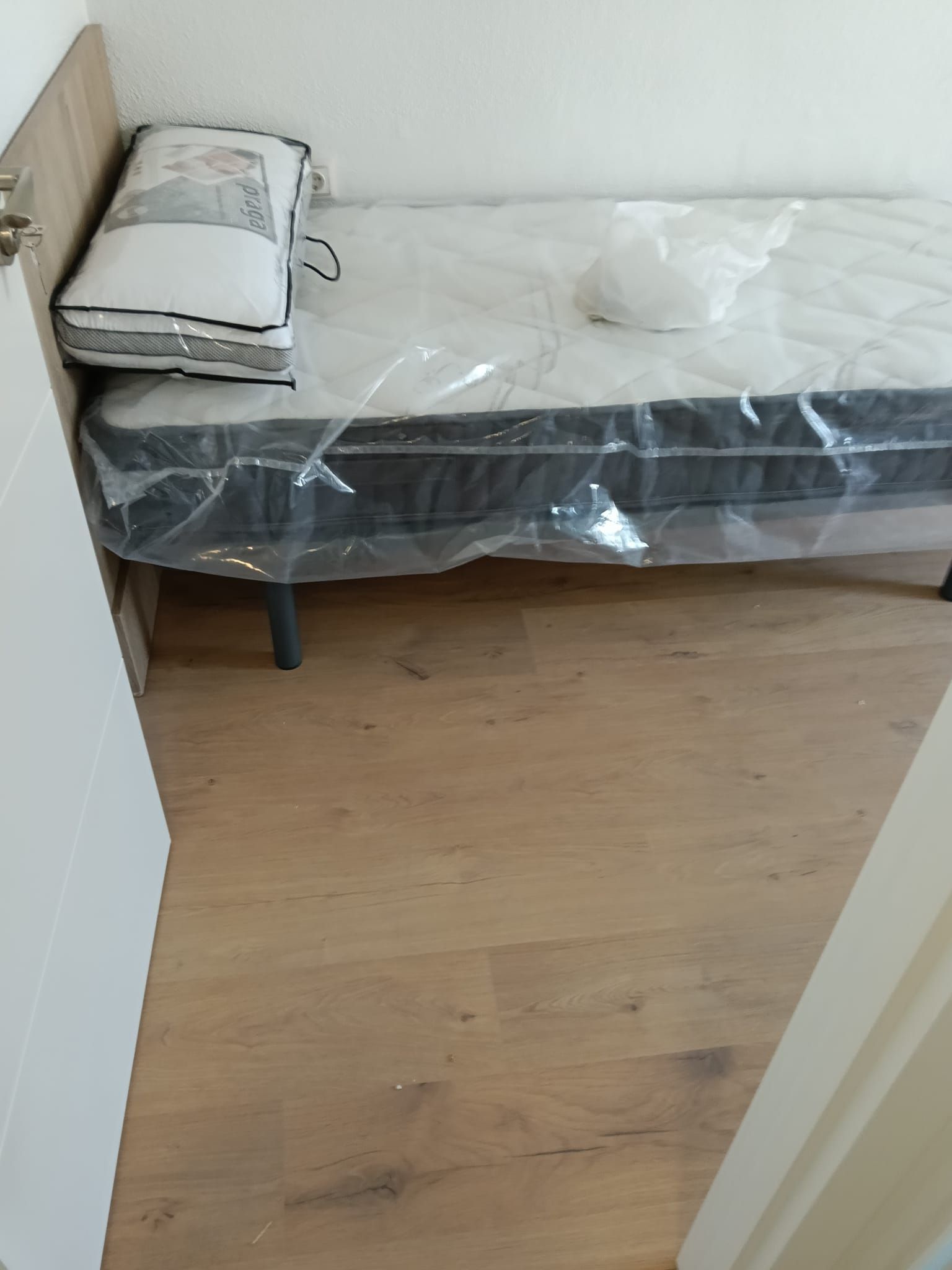 Dormitorio pequeño amueblado con una cama individual cubierta de plástico y ropa de cama doblada en el cabecero.