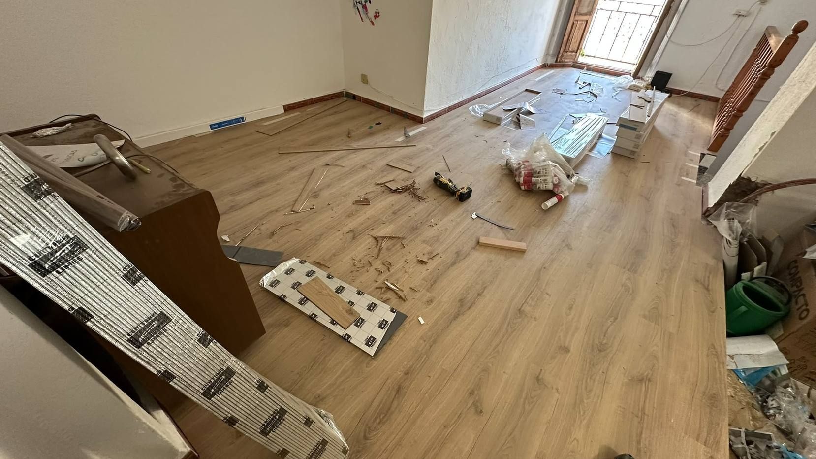 Habitación desordenada con suelo de madera, escombros dispersos, cajas y herramientas junto a una ventana y unas escaleras