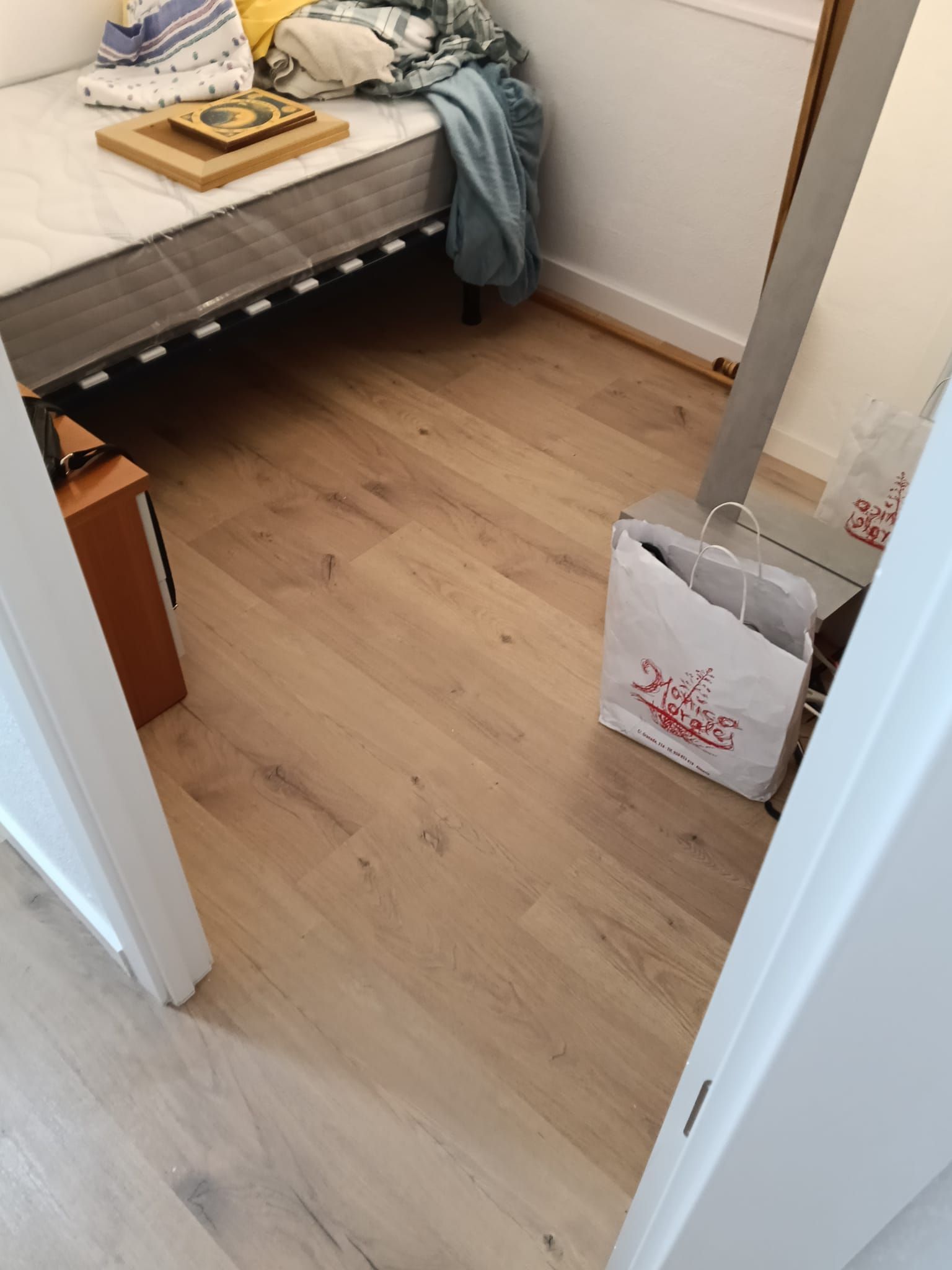 Vista de un dormitorio pequeño desde la puerta, con una cama, suelo de madera y una bolsa blanca de compras junto al espejo.
