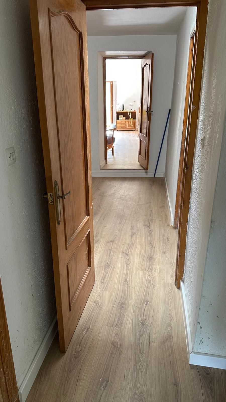 Pasillo estrecho con puertas de madera abiertas, paredes blancas y suelo de madera clara que conduce a una habitación luminosa.